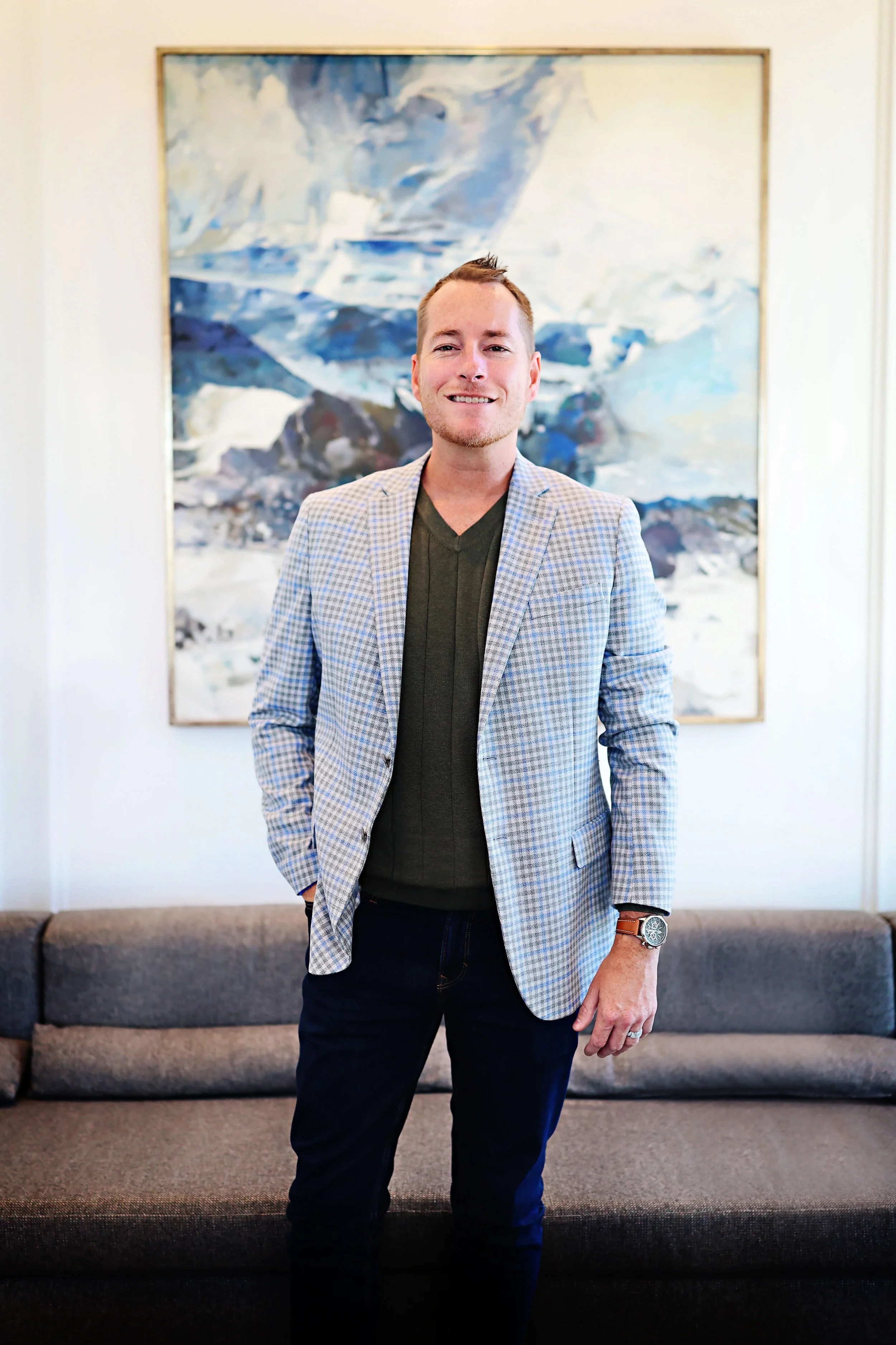 A man standing indoors in front of an abstract painting, wearing a checked blazer, dark shirt, black pants, a watch, and a ring, smiling at the camera.