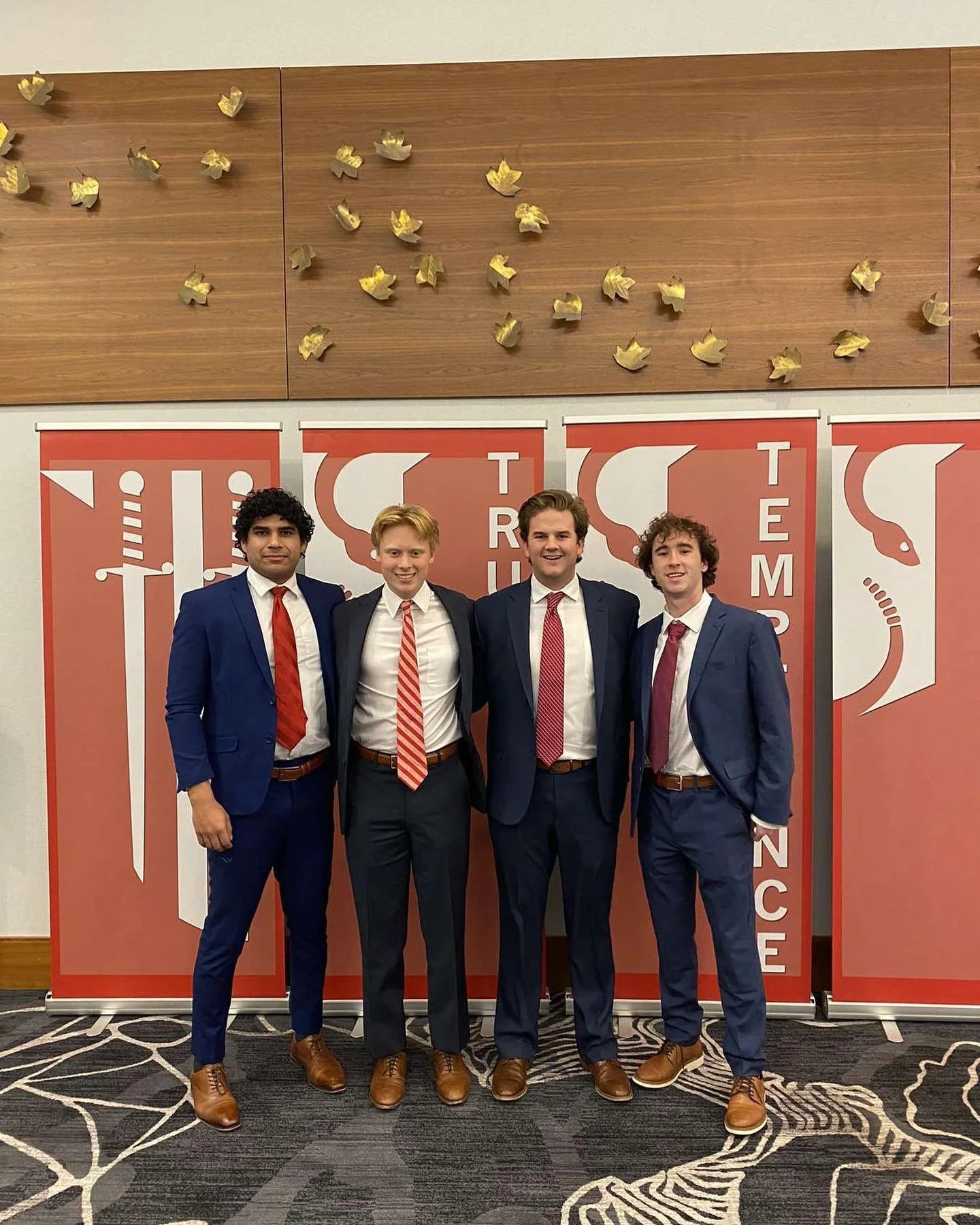 Four young men dressed in suits and ties standing side by side in front of a red and white banner with the words "Tri" and "Finance" visible, inside a room with patterned carpet and wooden wall with decorative gold leaves.