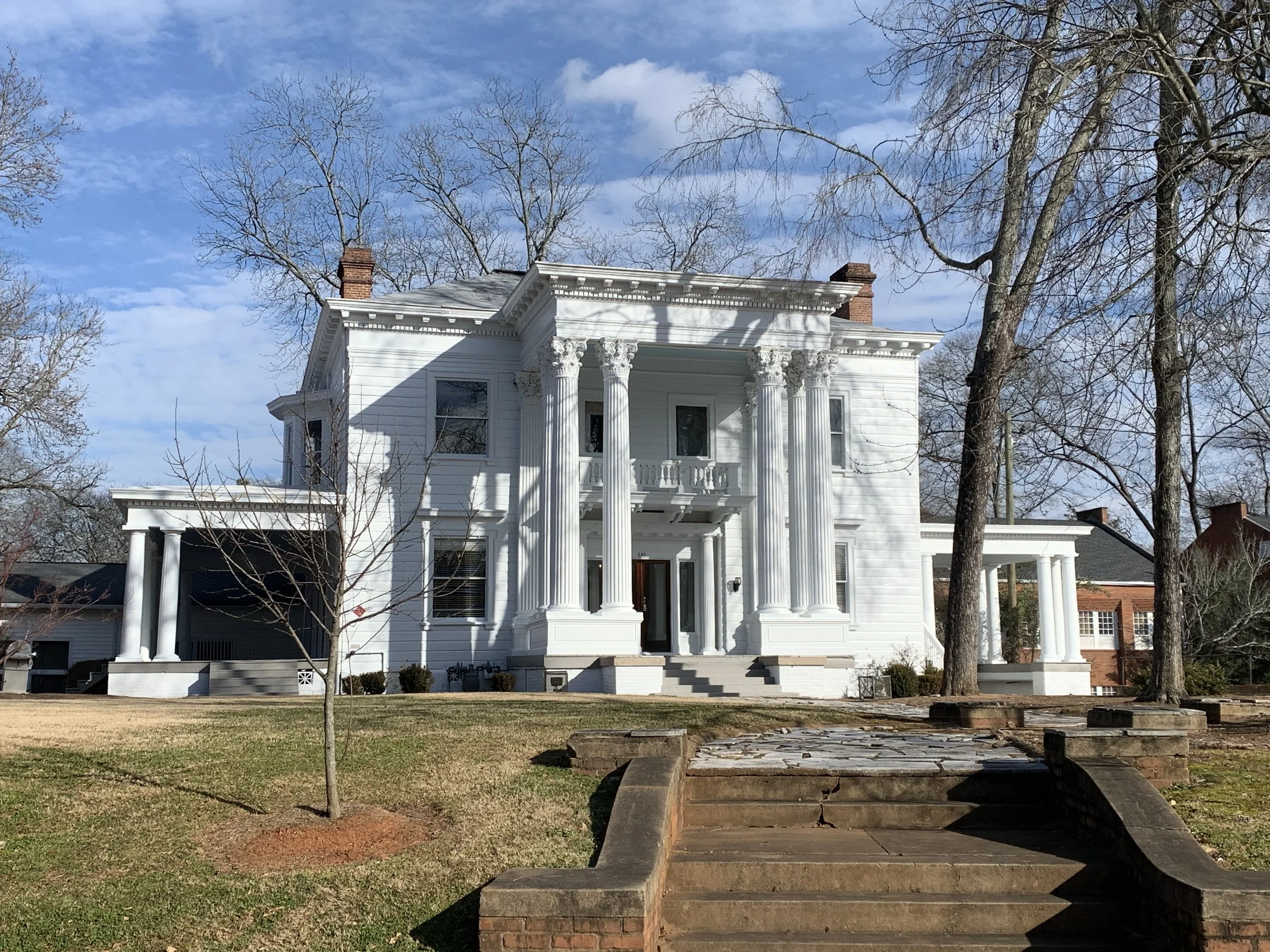 White historic house with large columns in front, stairs leading up to the entrance, surrounded by leafless trees, under a blue sky with some clouds.