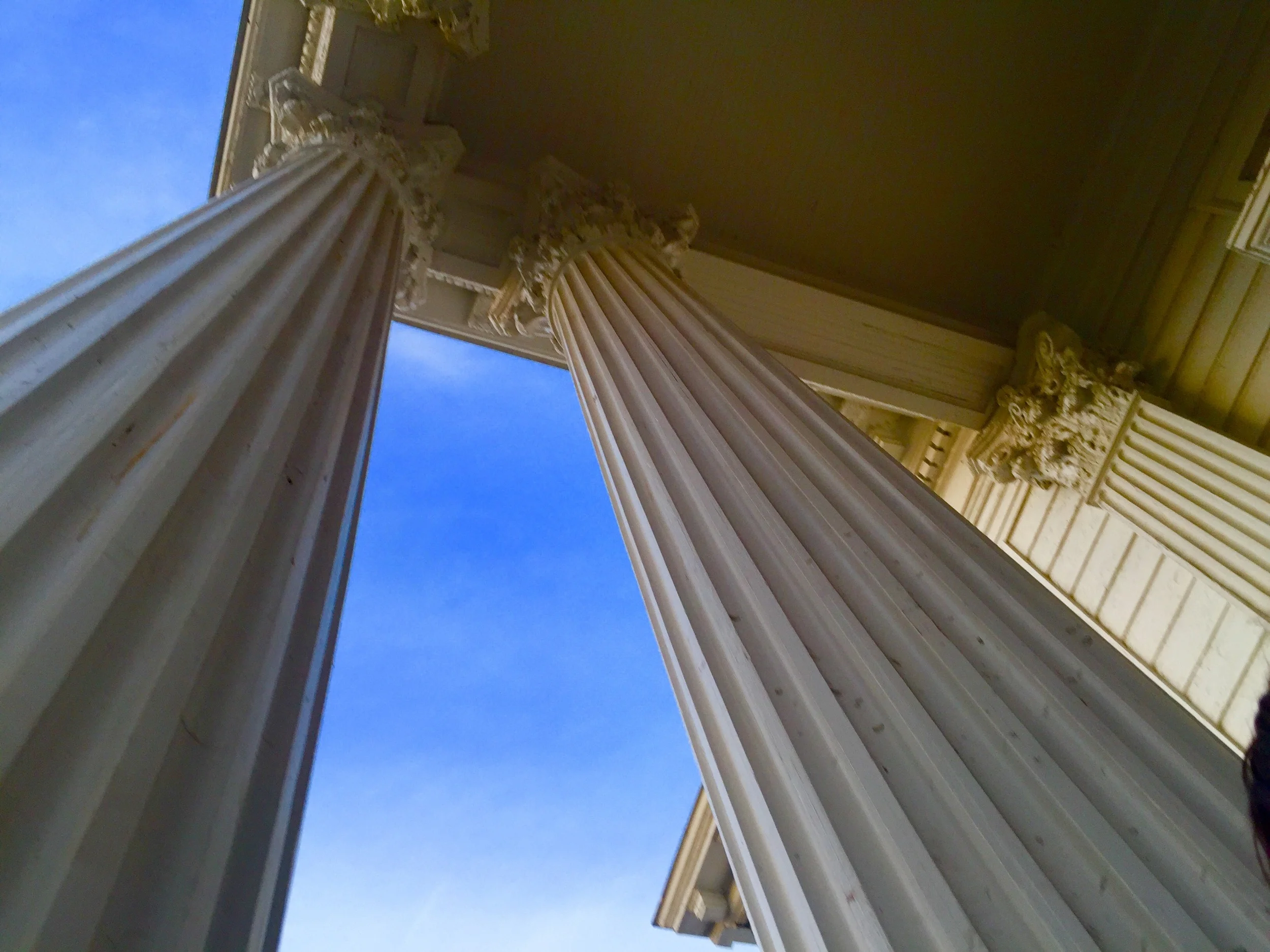 Looking up at two large, ornate, white classical columns with intricate capitals supporting a porch roof, against a bright blue sky.