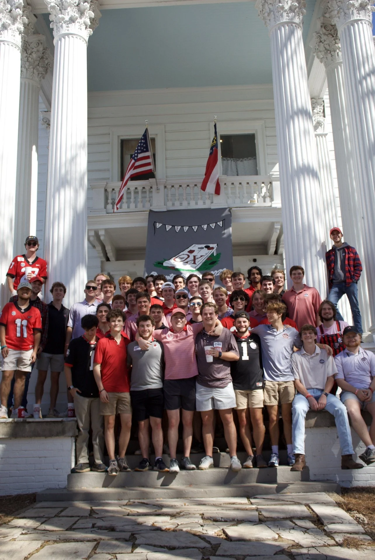 Group of young men on the front steps of a white house with large columns, American flags, and a banner with a poker theme.