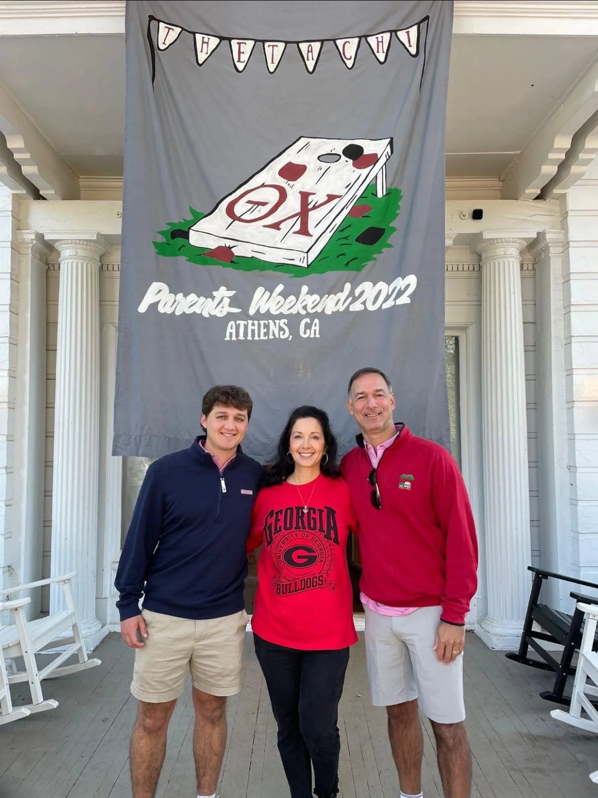 Three people standing in front of a large banner for a parents' weekend event at Athens, Georgia in 2012. The banner features an image of a cornhole game and text reading 'I VET A CH' and 'Parents Weekend 2012 Athens, GA'. The group includes a young man, a woman, and an older man, all smiling and dressed casually.