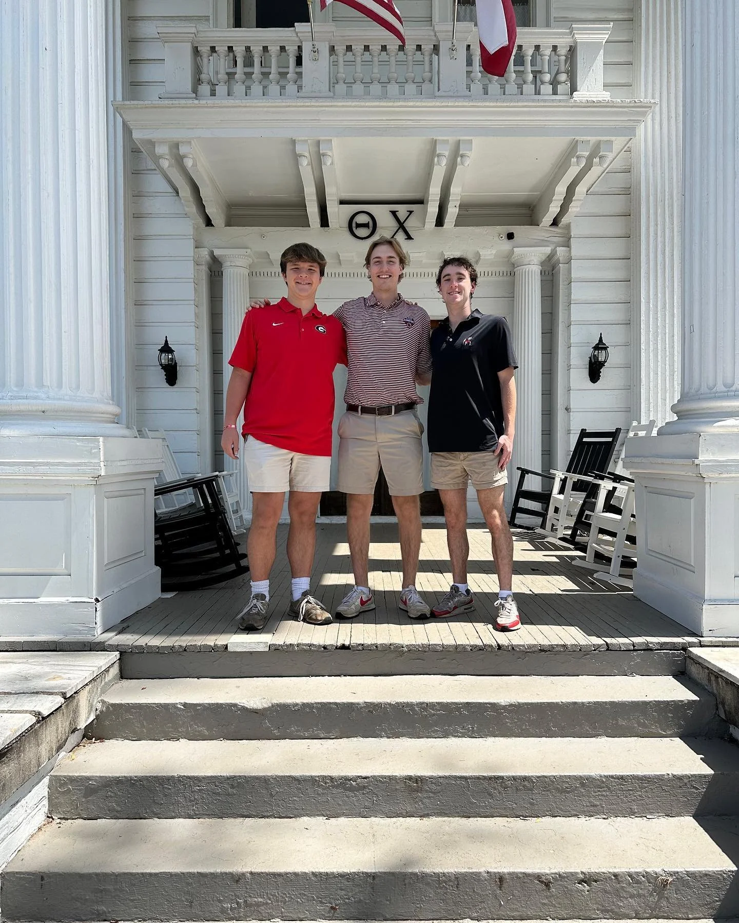 Three young men standing together on a porch in front of a white building with columns, American flags, and Greek letters, smiling at the camera.