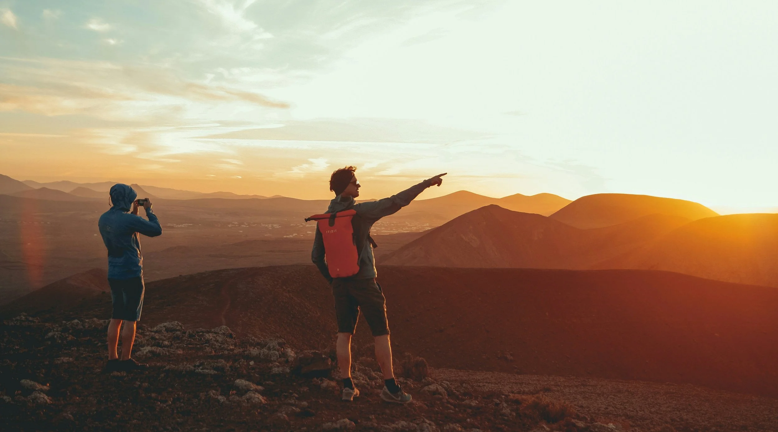 Two hikers on a mountain at sunset, one taking a photo and the other pointing towards the horizon.