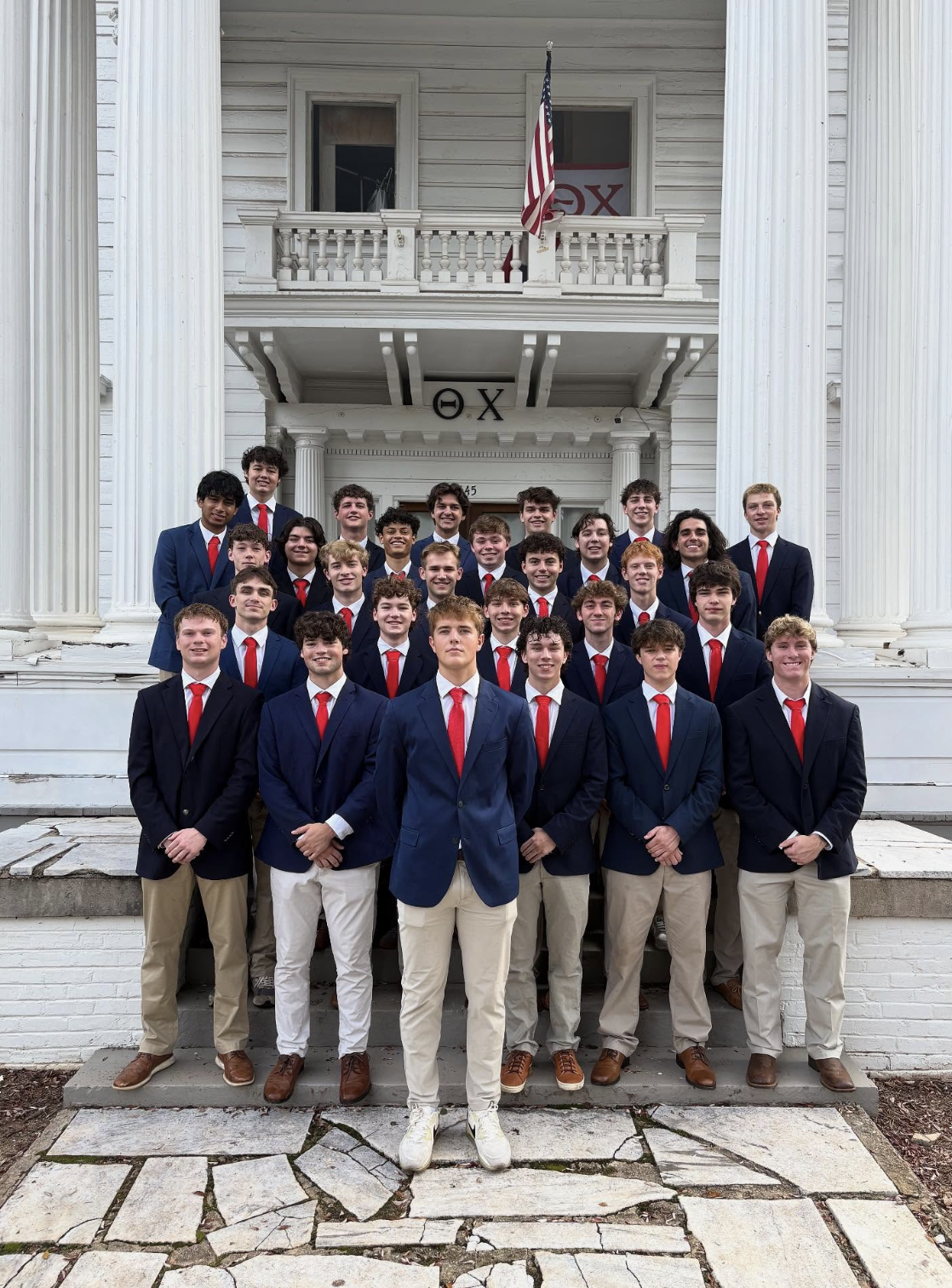Group of young men in formal wear with navy blazers, red ties, and khaki or beige pants, standing on the steps of a white building with classic columns and an American flag. It appears to be a graduation or fraternity photo.