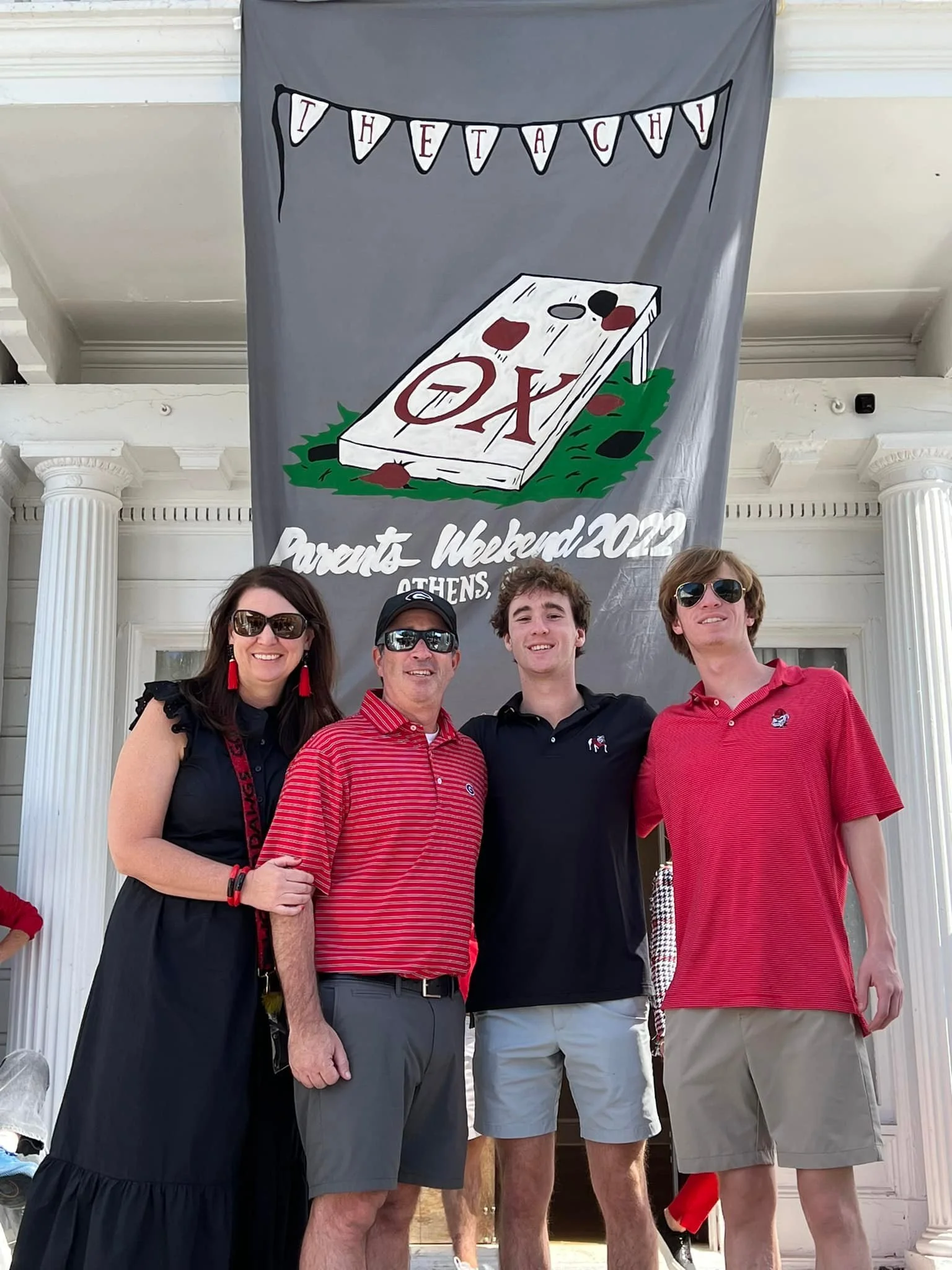 Family of four smiling and posing in front of a large banner with a giant tic-tac-toe game and the words "Parent Weekend 2022 Athens". The family members are wearing red and black clothing and sunglasses.