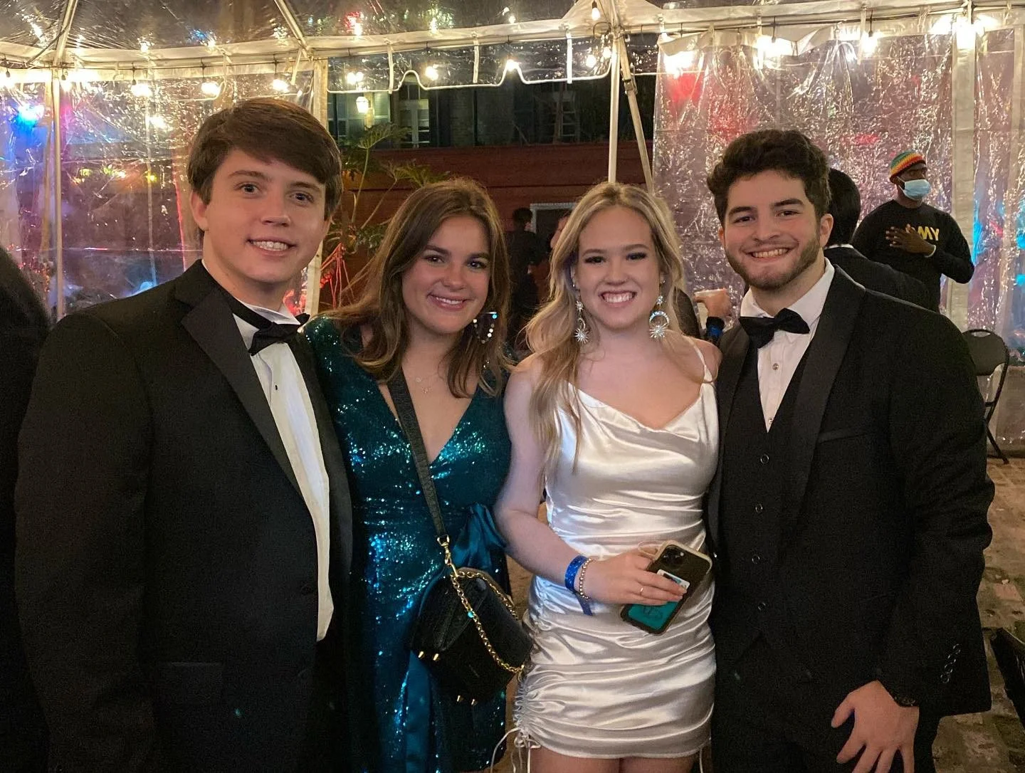 Four young adults dressed in formal attire standing together at a celebration event, with a decorated tent and a person wearing a mask in the background.