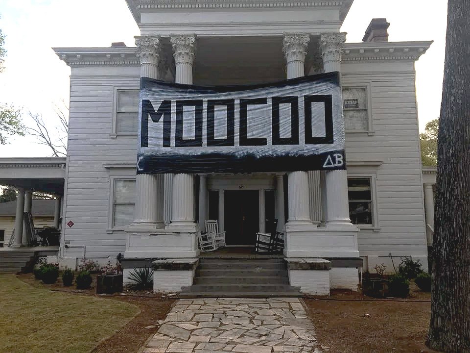 An old white house with columns, a banner hangs on the front reading 'MOODOO', and there are chairs and plants in front.