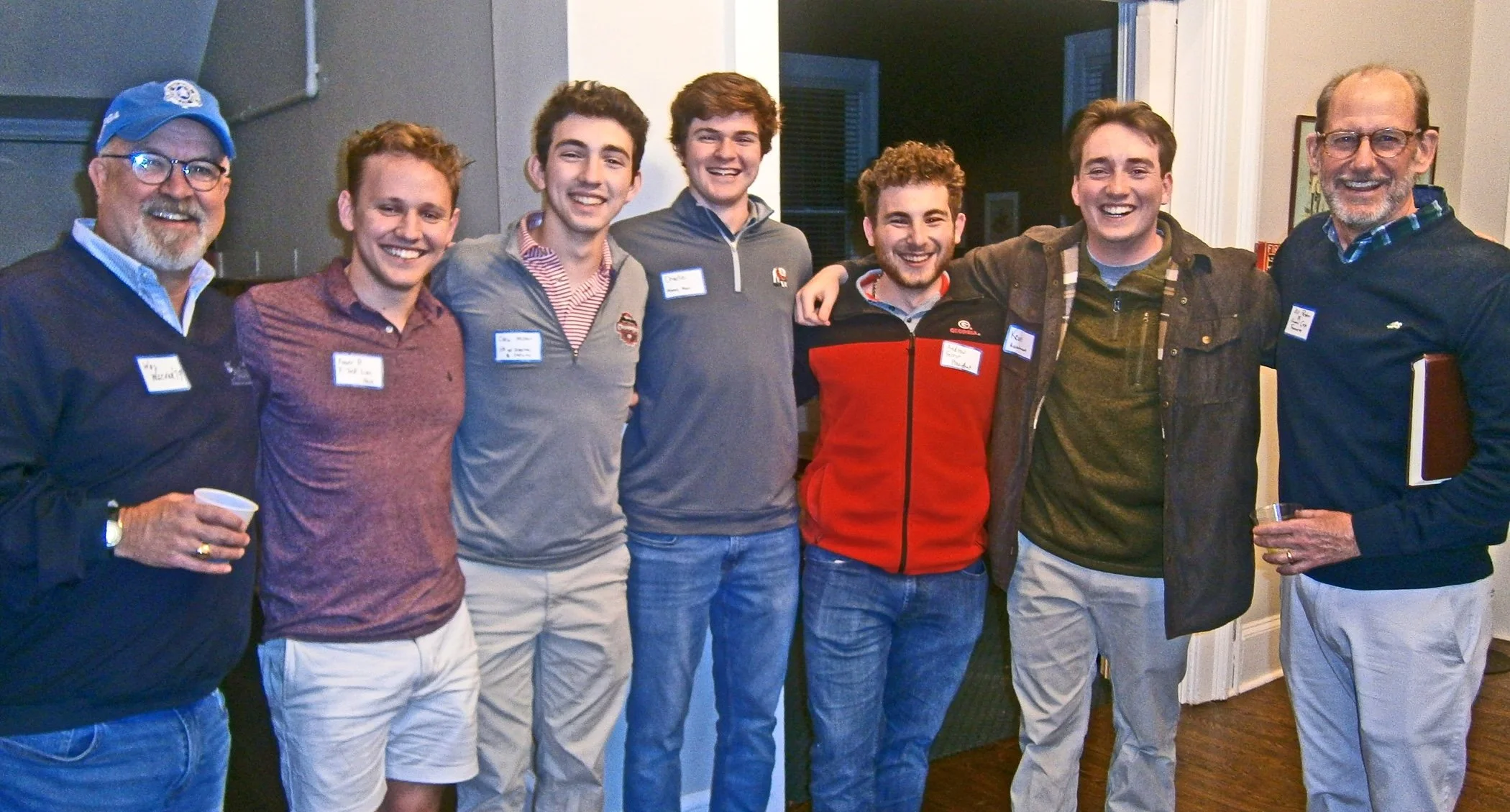 Group of seven men standing arm-in-arm indoors, smiling and posing for a photo at a social gathering.