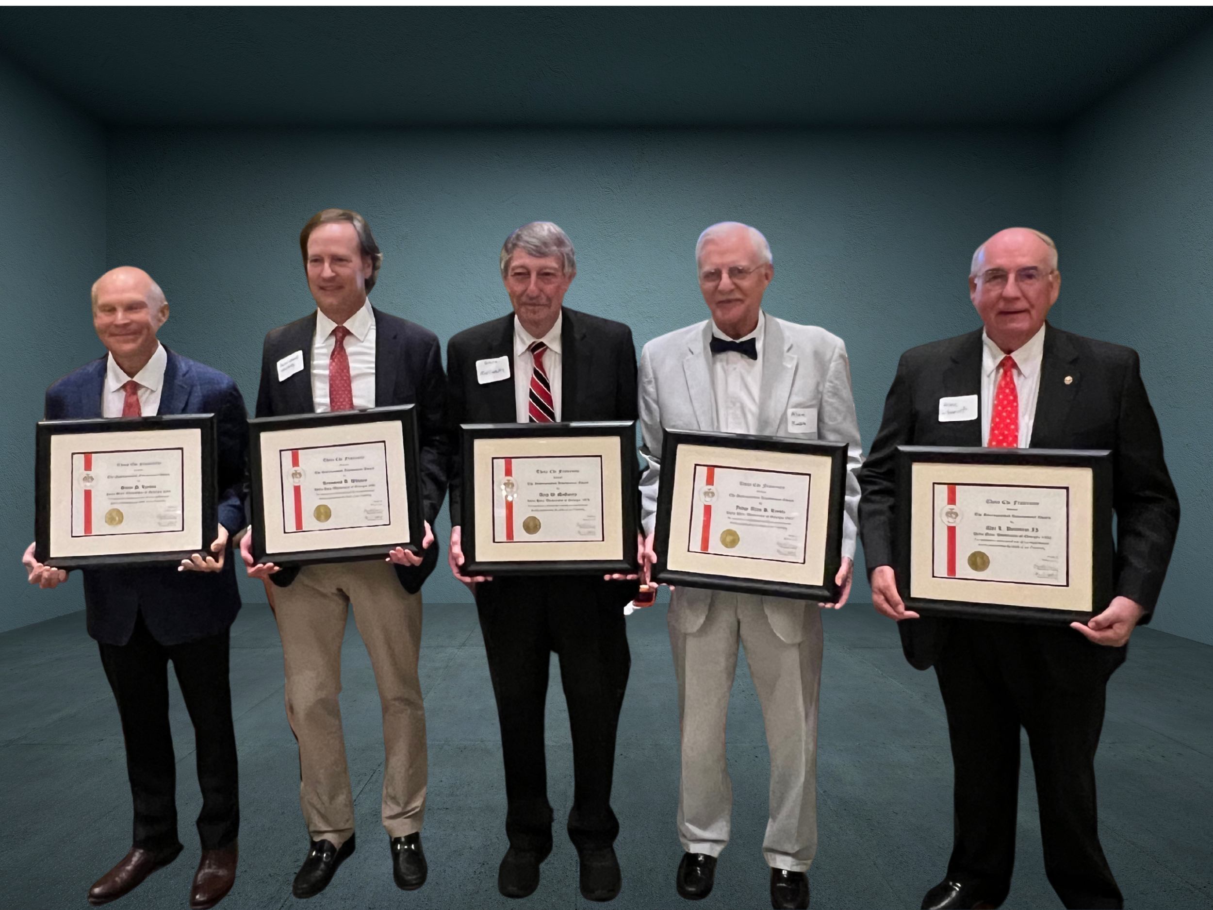 Five men in formal attire standing in a line, each holding framed certificates, inside a plain room with teal-colored walls.
