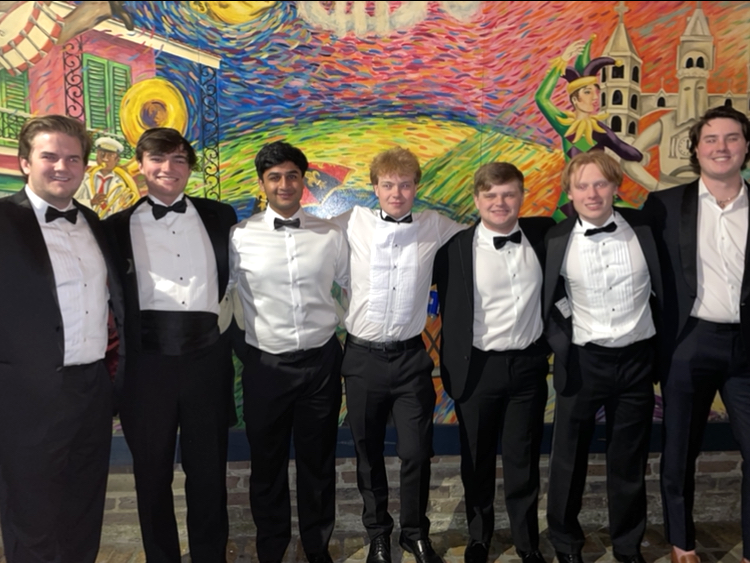 Seven young men dressed in tuxedos with bow ties posing in front of a colorful mural of a castle and a figure with a hat, sword, and cape.
