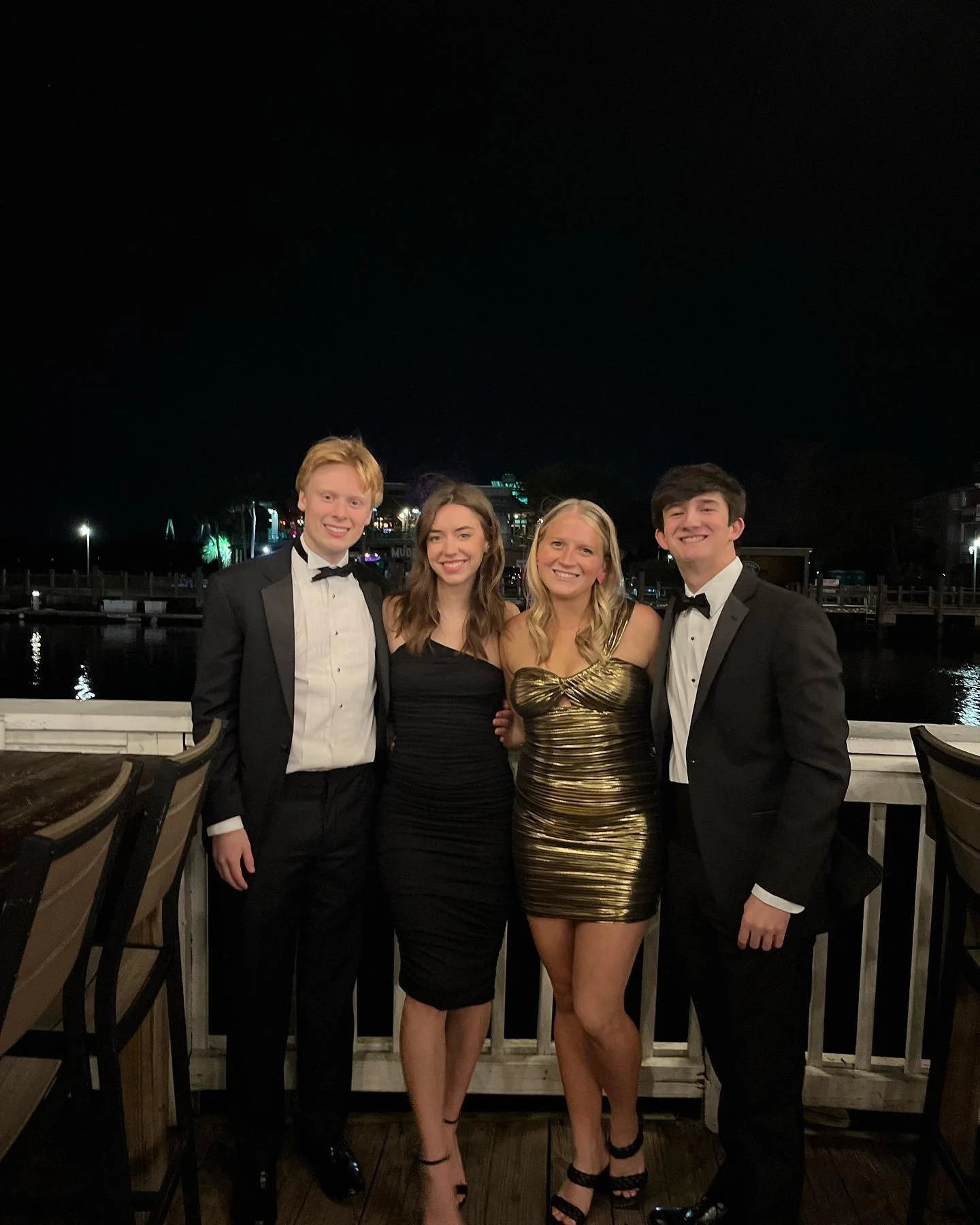 Four young adults dressed in formal attire at night, standing on a wooden deck near water, with city lights in the background.