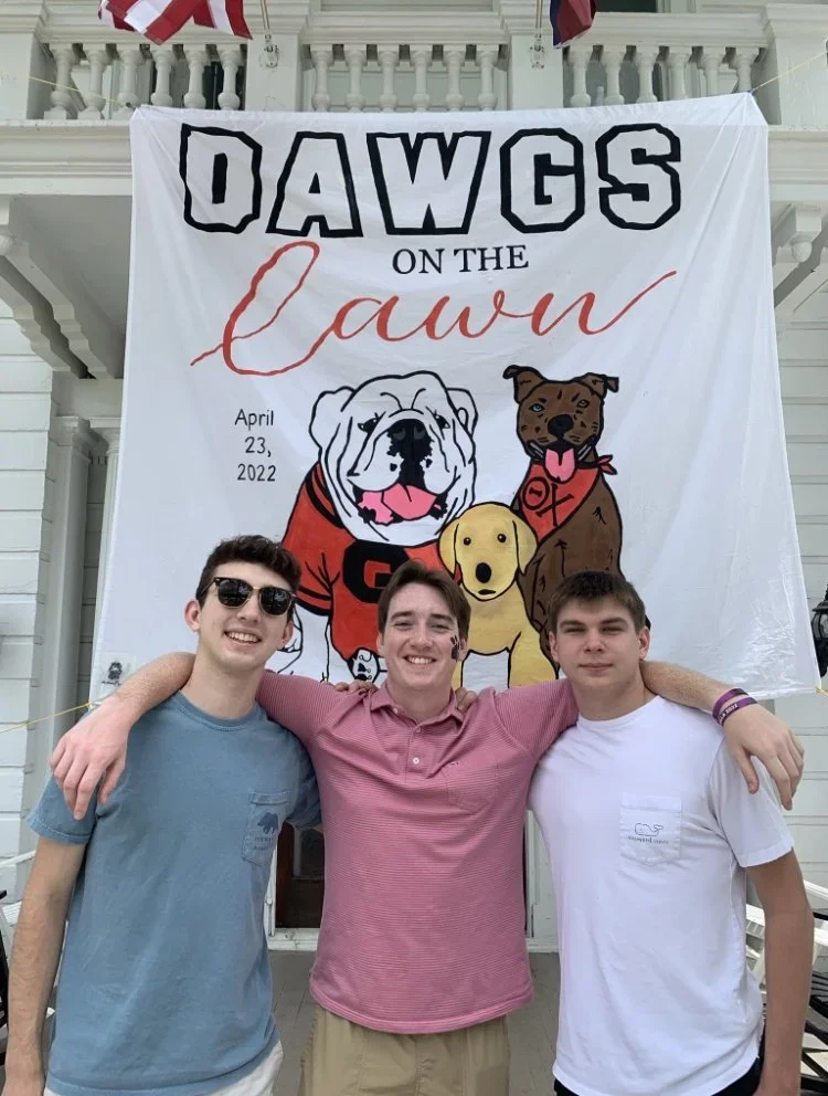 Three young men stand in front of a large banner with cartoon dogs and the words 'Dawgs on the Cawn,' dated April 23, 2022. They are smiling and have their arms around each other.