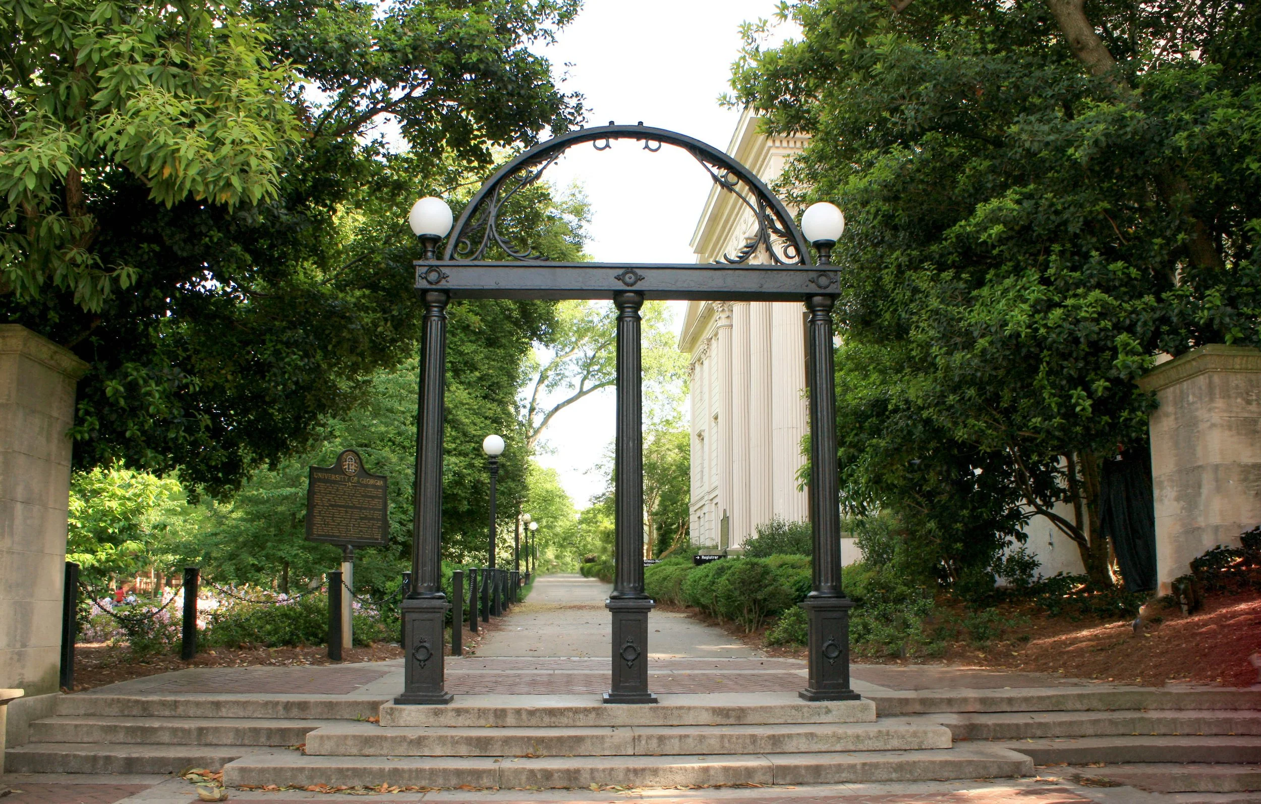 Entrance gate to the University of Georgia, with a decorative black archway and white globe lights, flanked by trees and a sidewalk.