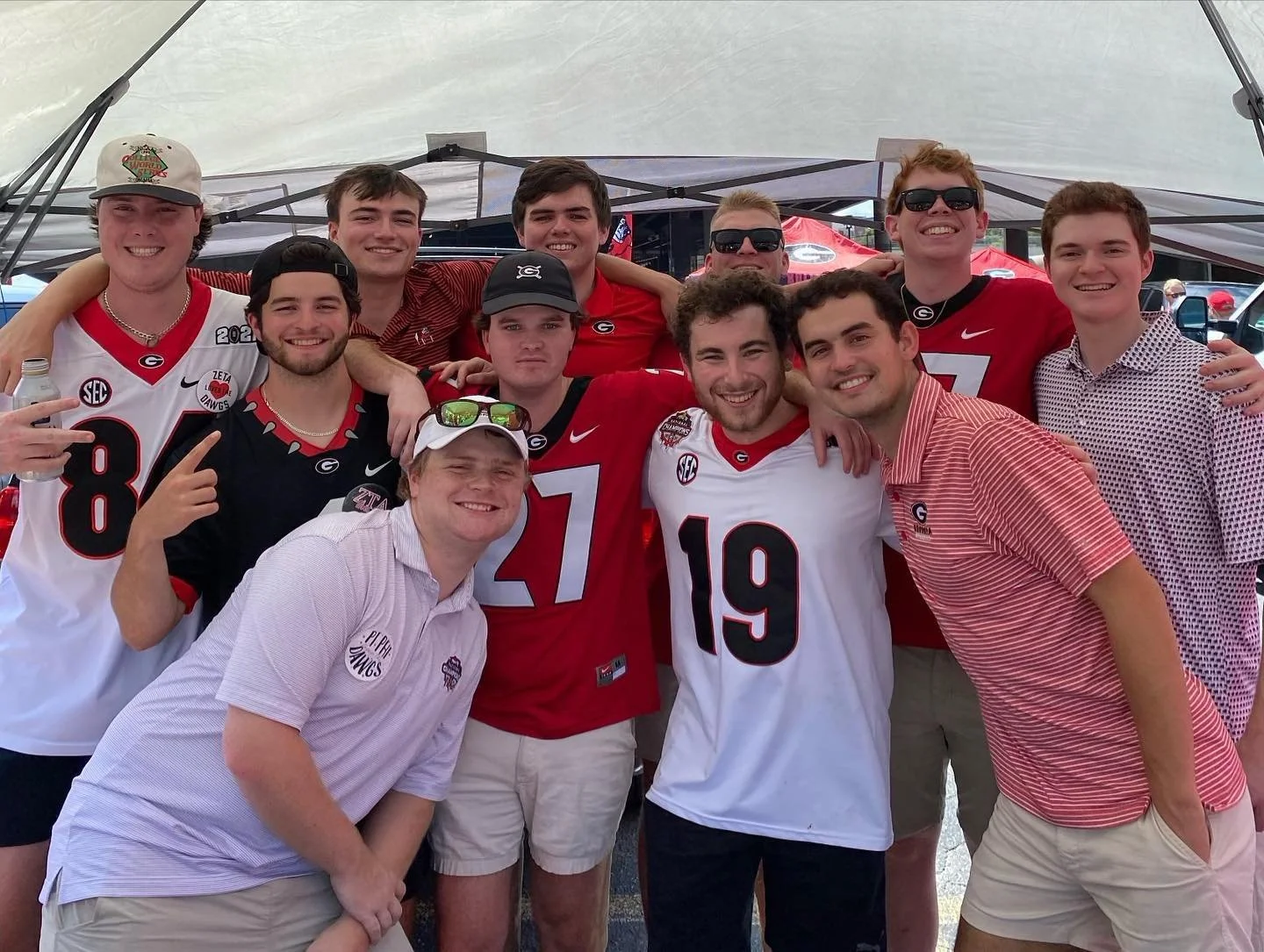 Group of young men wearing University of Georgia football jerseys and casual clothing, standing and smiling inside a large tent at a tailgate or sports event.