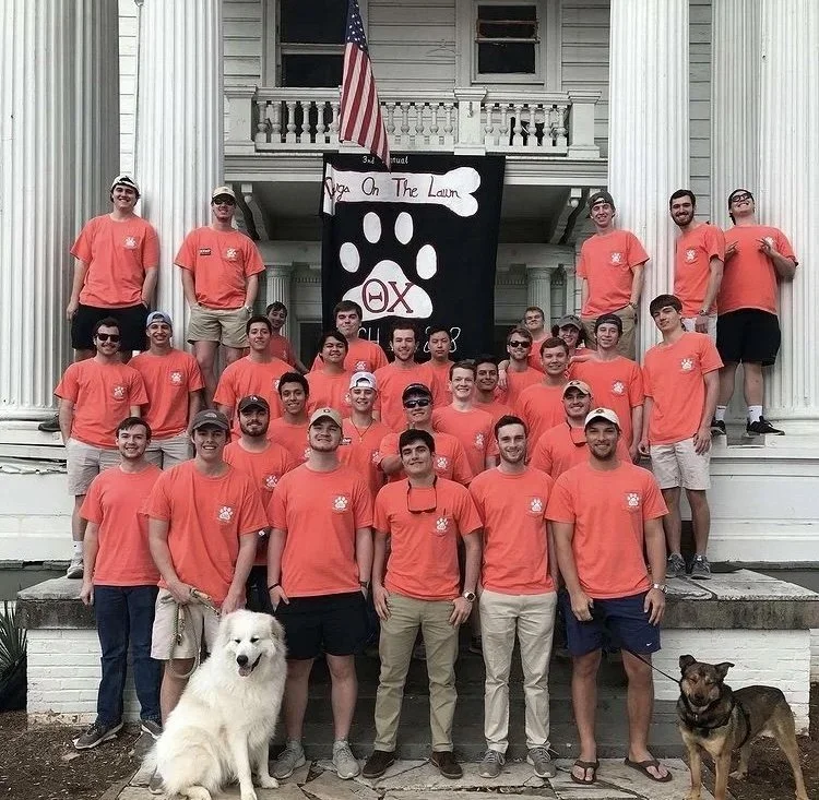 A group of men and women wearing bright orange T-shirts standing on the steps of a large white building, with some sitting and others standing. There are two dogs in the front, one large white fluffy dog and one smaller brown and black dog. A black banner with a Paw Print and Greek letters, along with a sign that says "Yaga on the Lawn," is displayed behind the group. The building has tall columns, a balcony, and an American flag hanging above the banner.