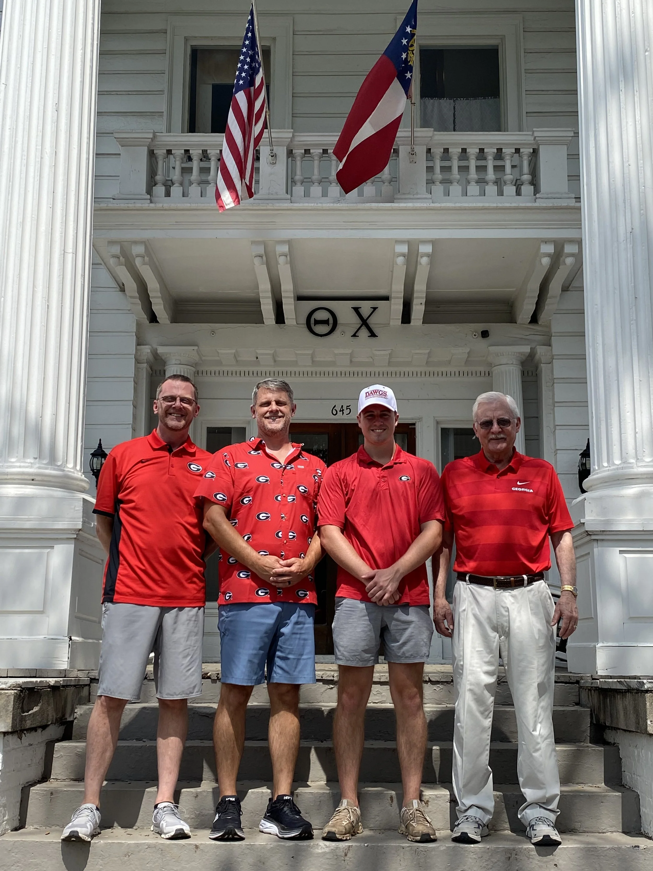 Four men standing on the steps of a white house with Greek letters above the door, wearing red shirts, with American and Georgia flags on the porch.