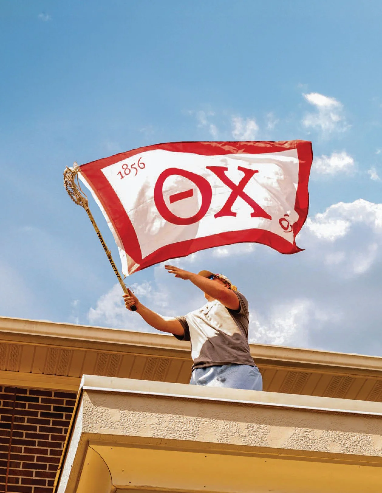 Person on a balcony waving a red and white flag with 'OX' and '1856' written on it against a blue sky with scattered clouds.