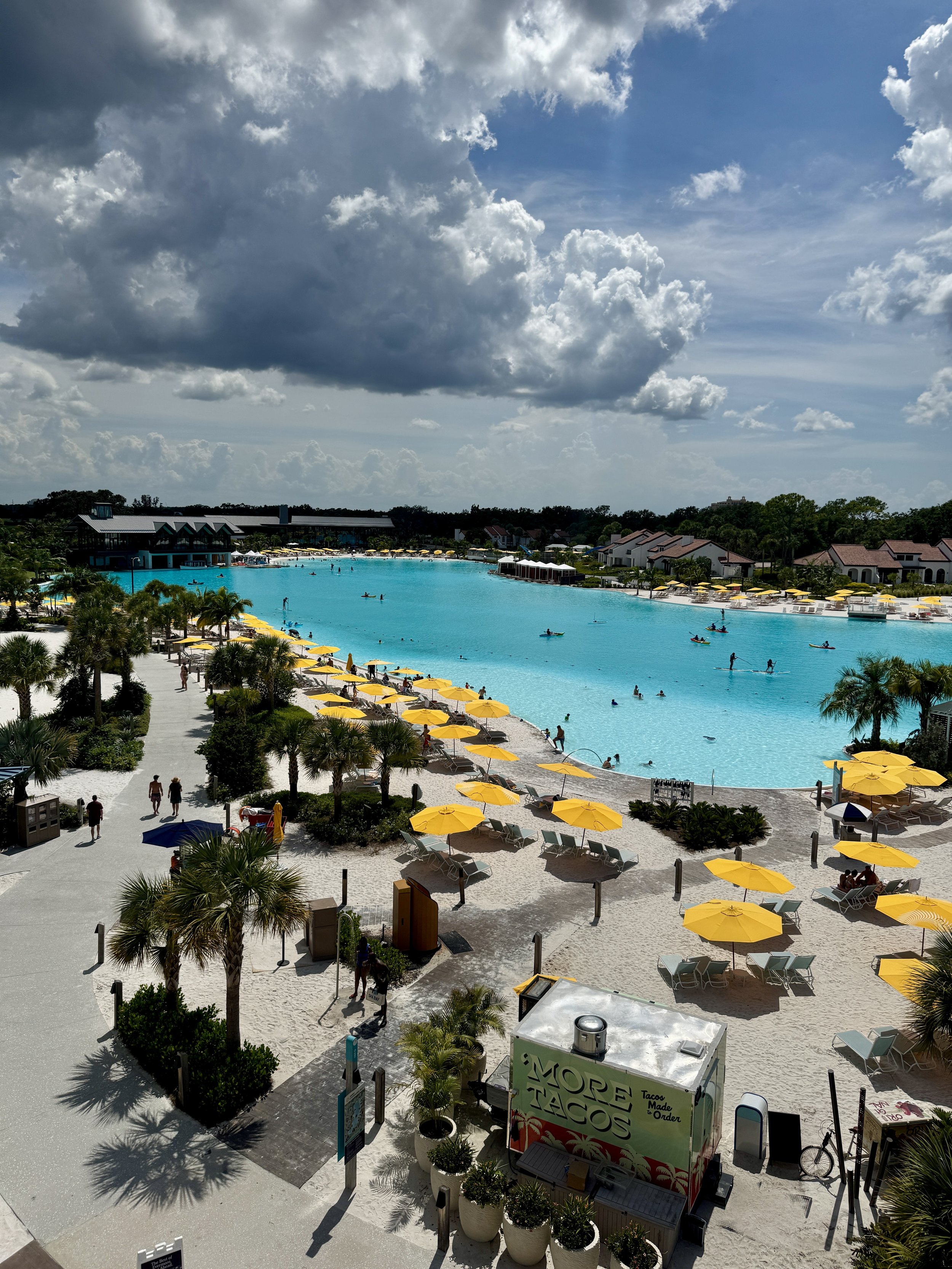 A large outdoor swimming pool with clear blue water, surrounded by yellow umbrellas and lounge chairs, under a cloudy sky, with people swimming and relaxing, and neighboring houses and palm trees nearby.