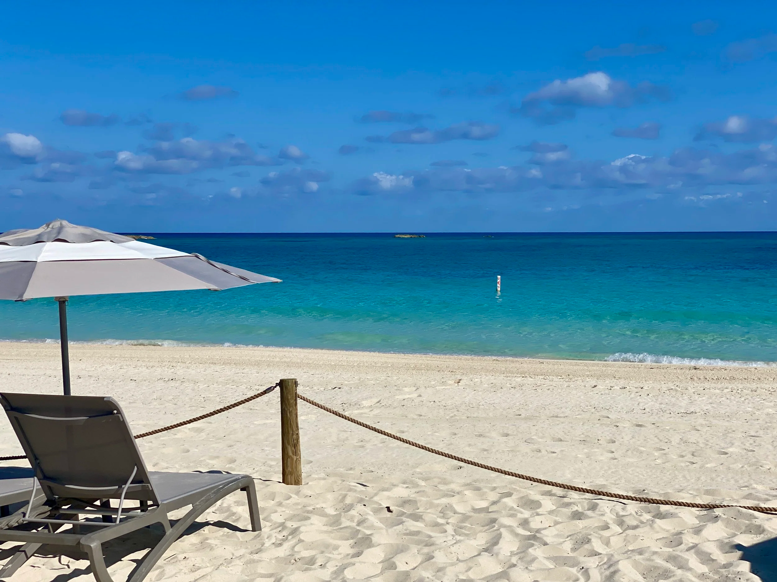 A beach with white sand, a gray lounge chair, a large white and gray umbrella, a rope barrier, and turquoise ocean water under a partly cloudy blue sky.