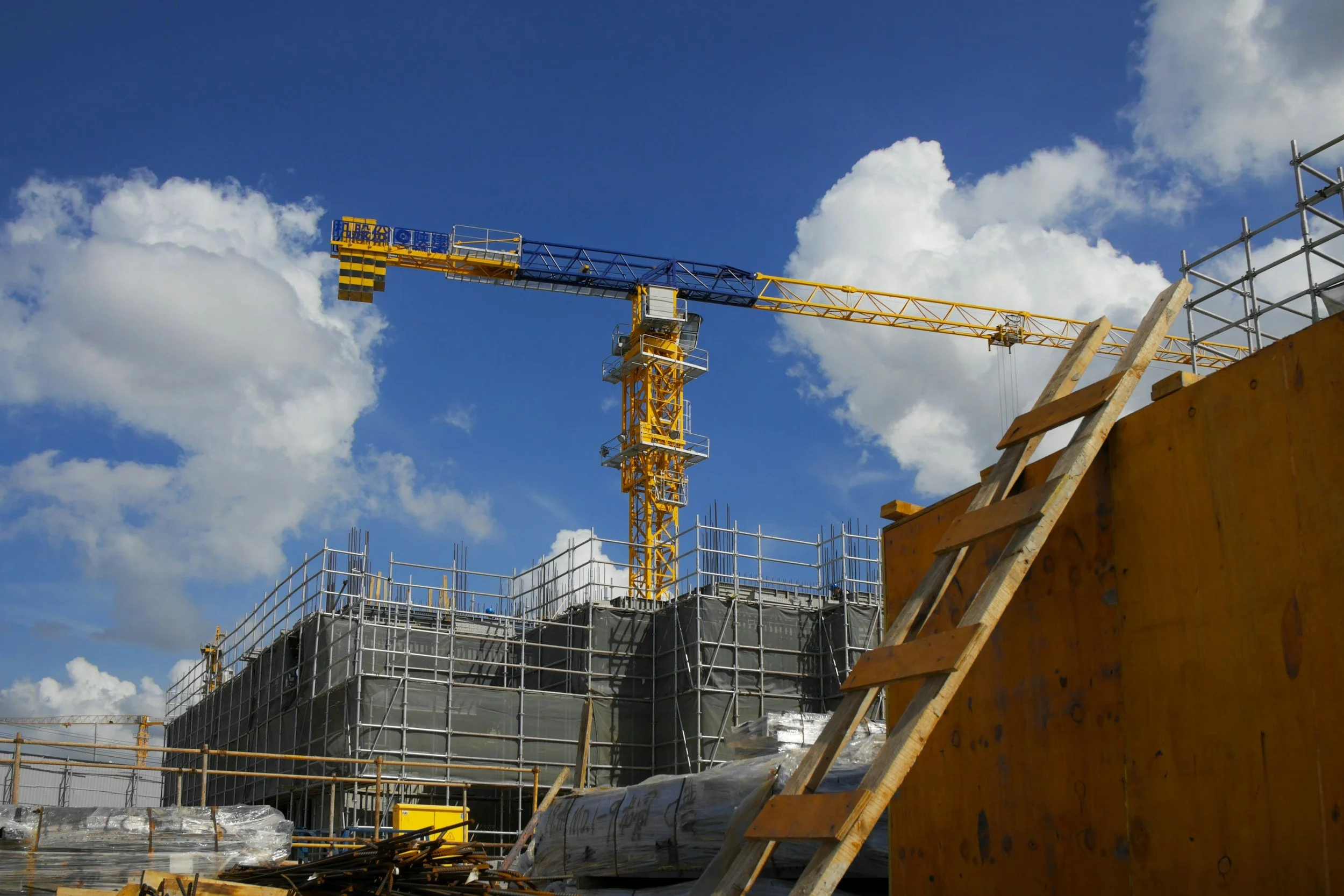 Construction site with a yellow tower crane, scaffolding around a building under construction, blue sky with clouds, wooden ladder leaning against a yellow partition, construction materials on the ground.