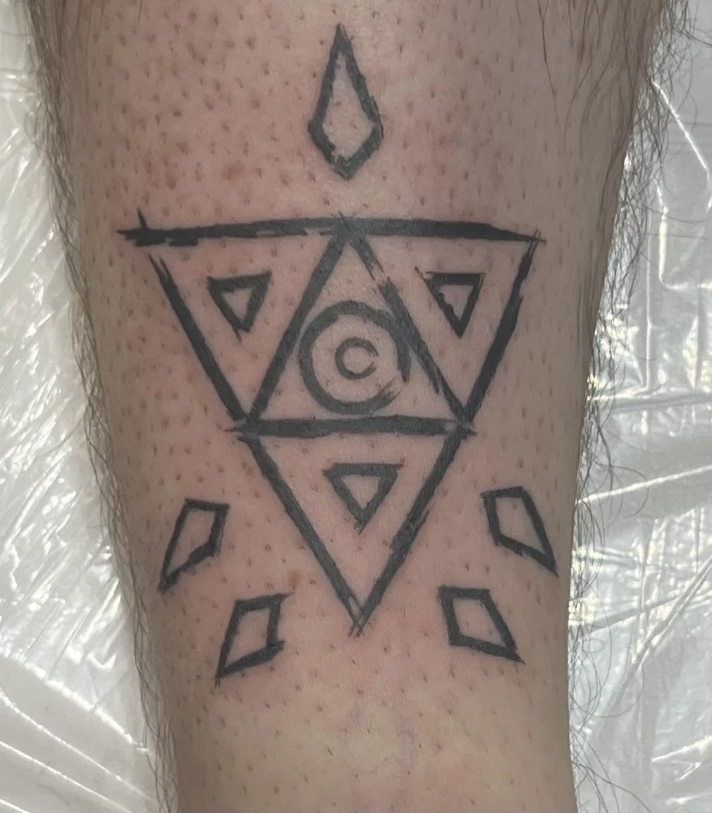 A fresh black ink tattoo on skin featuring geometric shapes including triangles, diamonds, and a circle with a smaller circle inside, arranged in a symmetrical pattern.