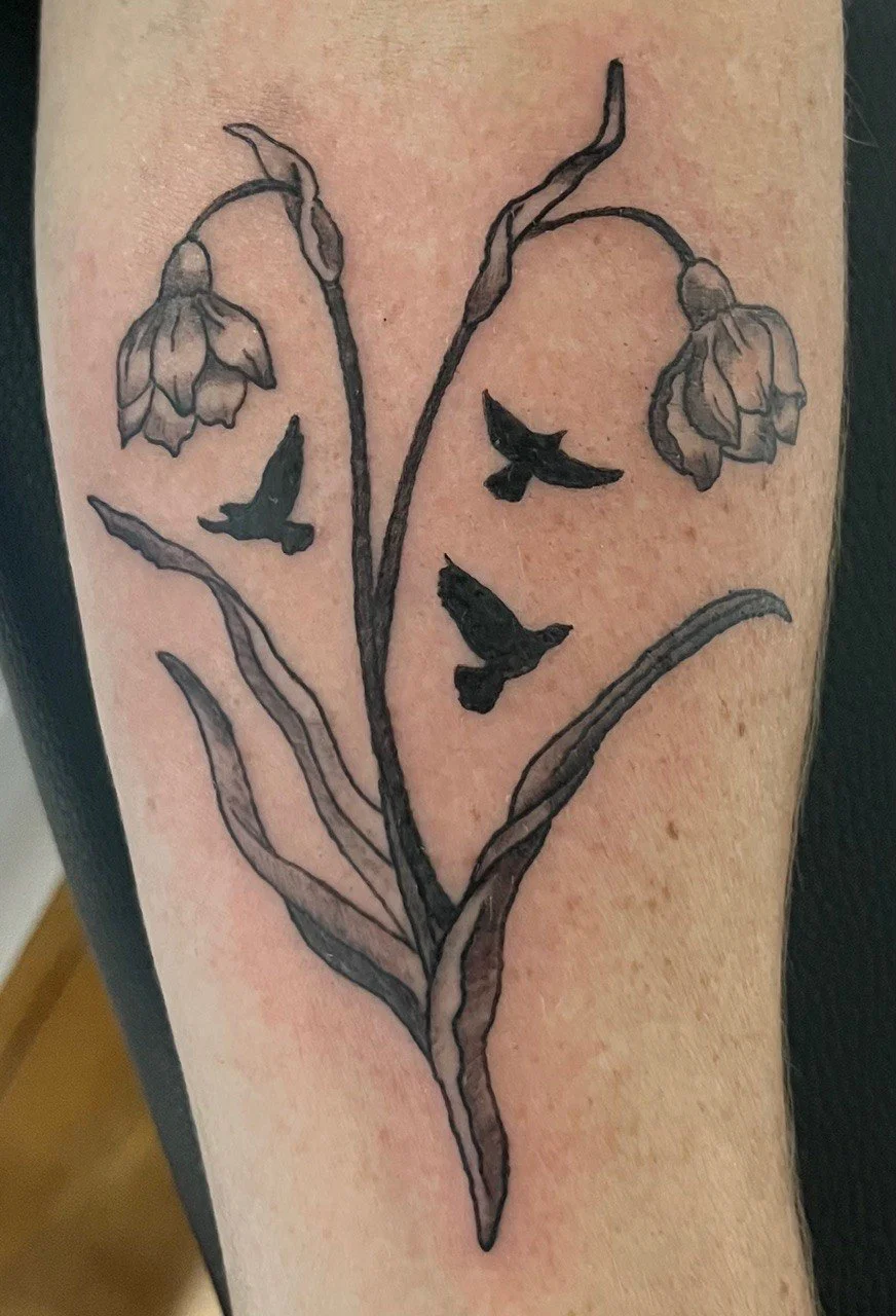 A black outline tattoo of a flower with long, curved leaves and three flying birds on a person's skin.