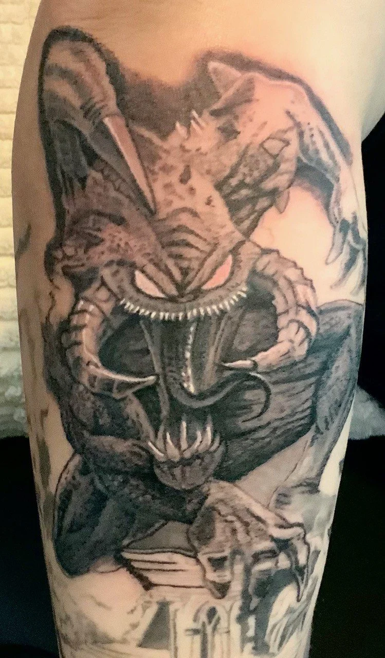 Tattoo of a fierce panther with wings, wearing a collar, perched on a stone pedestal with skulls, in black and gray ink.