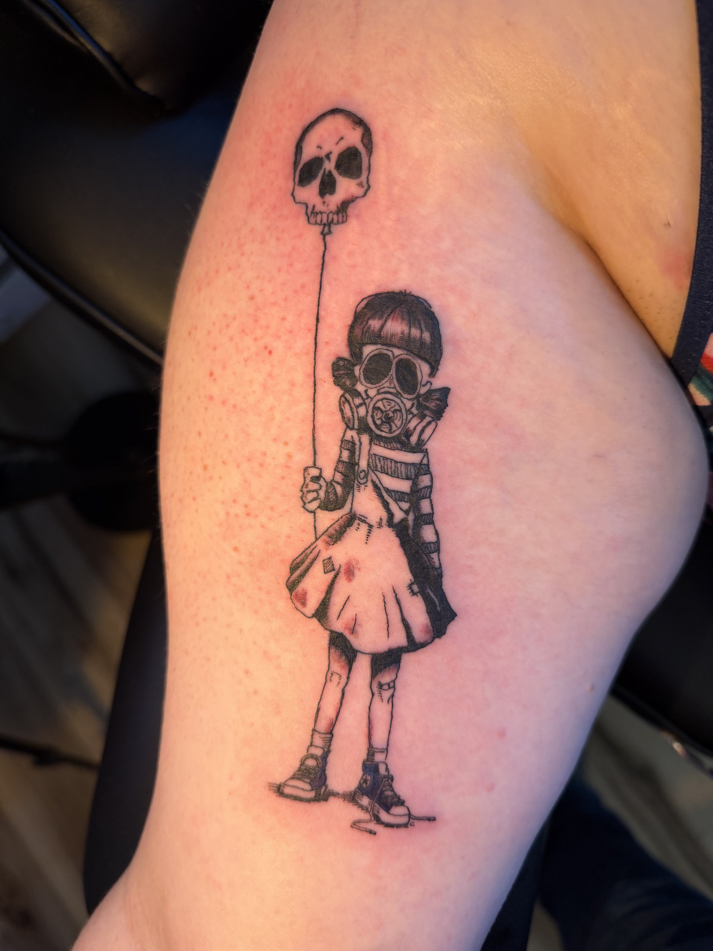 Tattoo of a girl wearing a gas mask and holding a balloon with a skull face, on the person's arm.