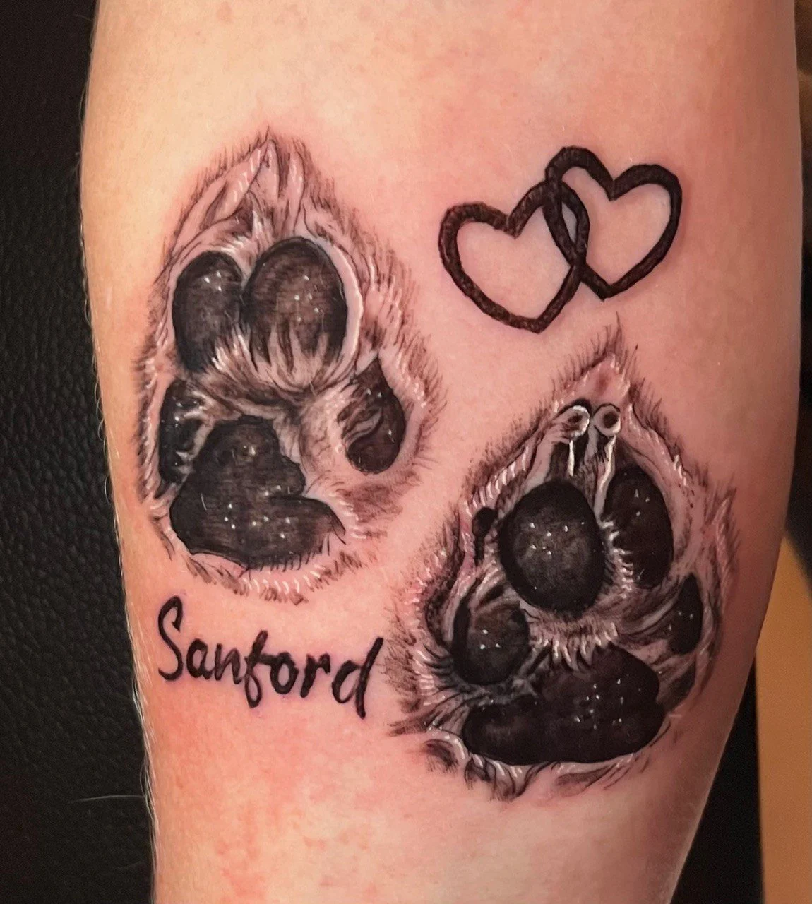 Tattoo of two realistic dog paw prints and two overlapping hearts above the paw prints. The word "Sandford" written below the paw prints.