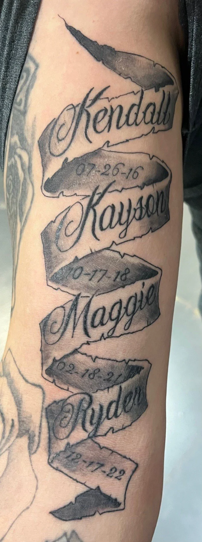 Tattoo with torn paper design featuring names Kendrick, Kayden, Maggie, Ryder, and dates 07-26-16, 10-17-18, 02-18-21, and 12-17-22.
