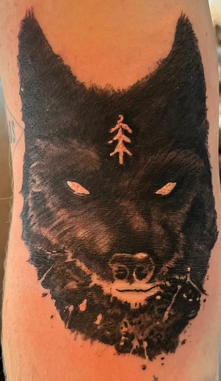 Tattoo of a black wolf's face on a person's arm, with white eyes and a small tree symbol on its forehead.