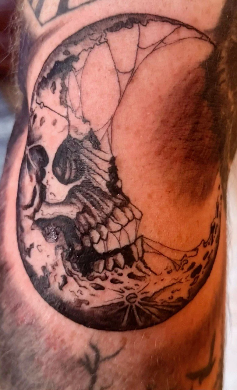 Black and gray tattoo of a human skull in profile with a circular design surrounding parts of the skull on a person's arm.
