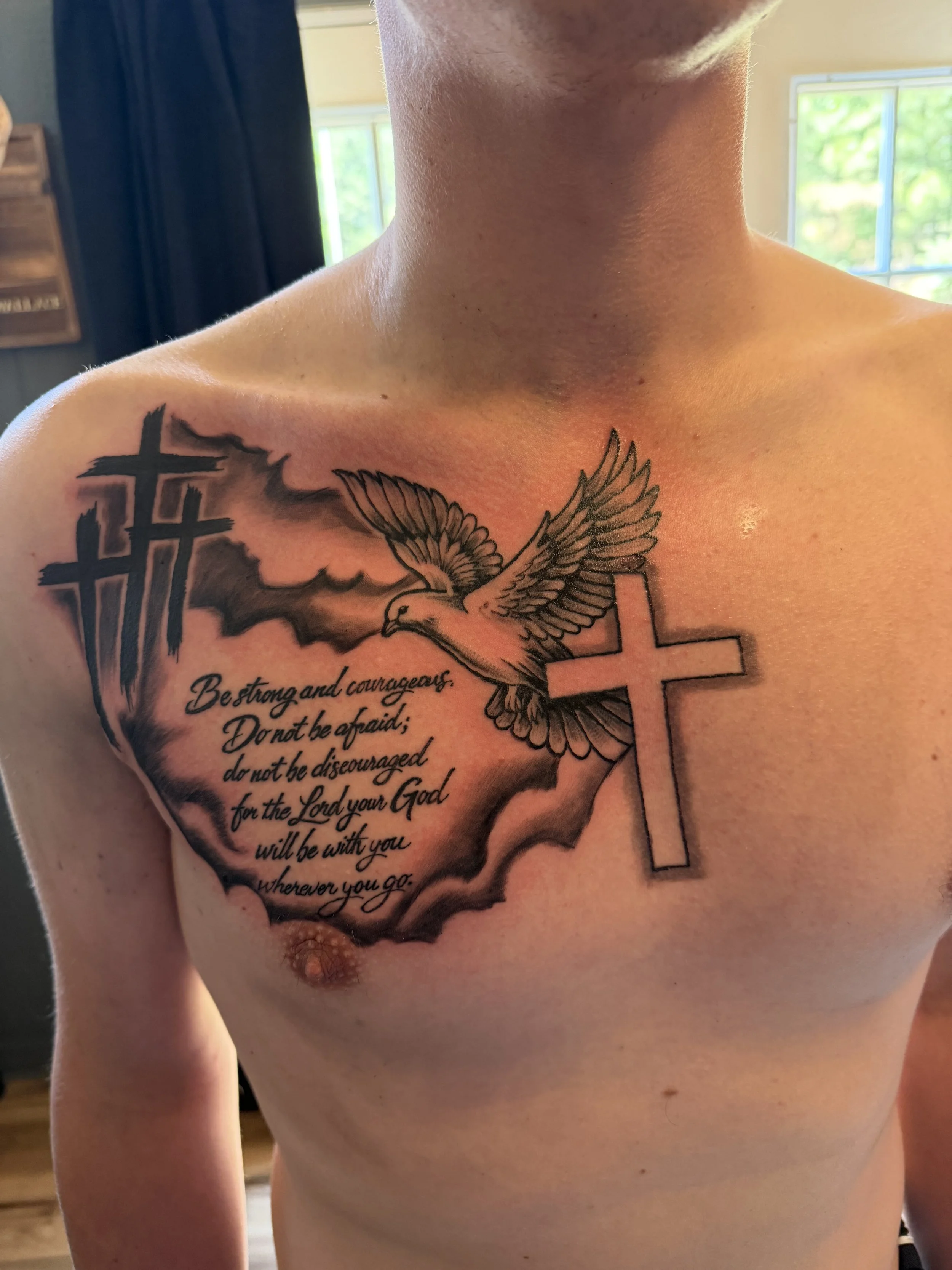 Tattoo on person's chest featuring a bird with wings spread, a cross, and a quote that reads "Be strong and courageous. Do not be afraid; do not be discouraged, for the Lord your God will be with you wherever you go."