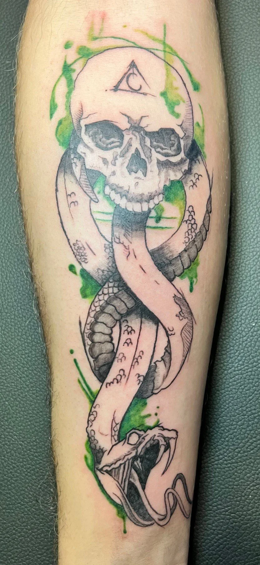 Tattoo of a skull with a serpent wrapped around it, including green ink splashes and an all-seeing eye inside a triangle on the forehead.