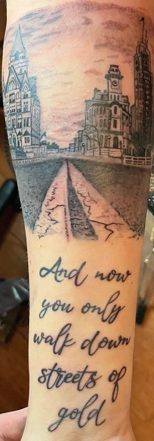 A tattoo of a cityscape with buildings and a street, along with a quote that reads, "And now you only walk down streets of gold."