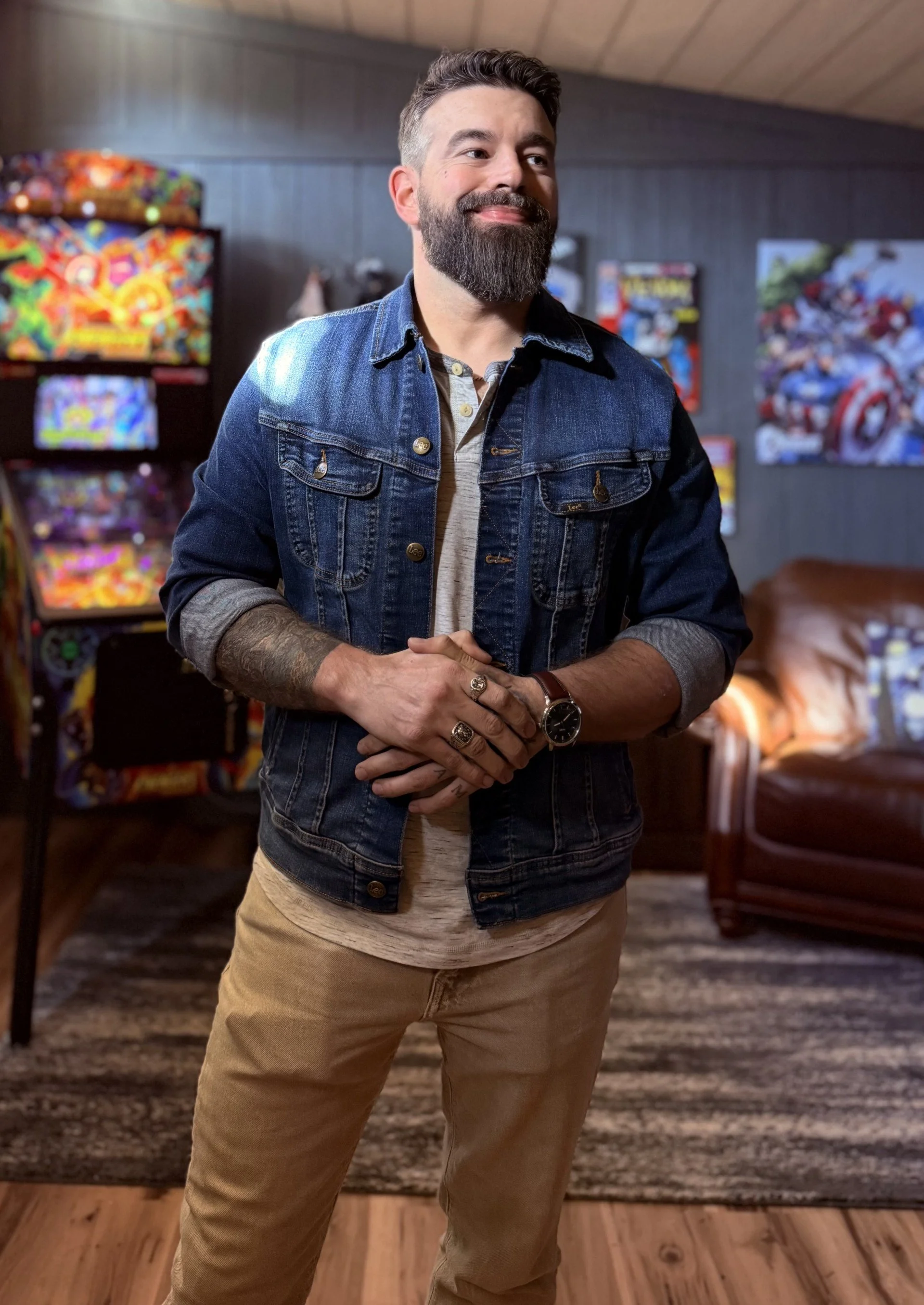 A man with a beard and tattooed arm standing in a room with pinball machines and posters on the wall