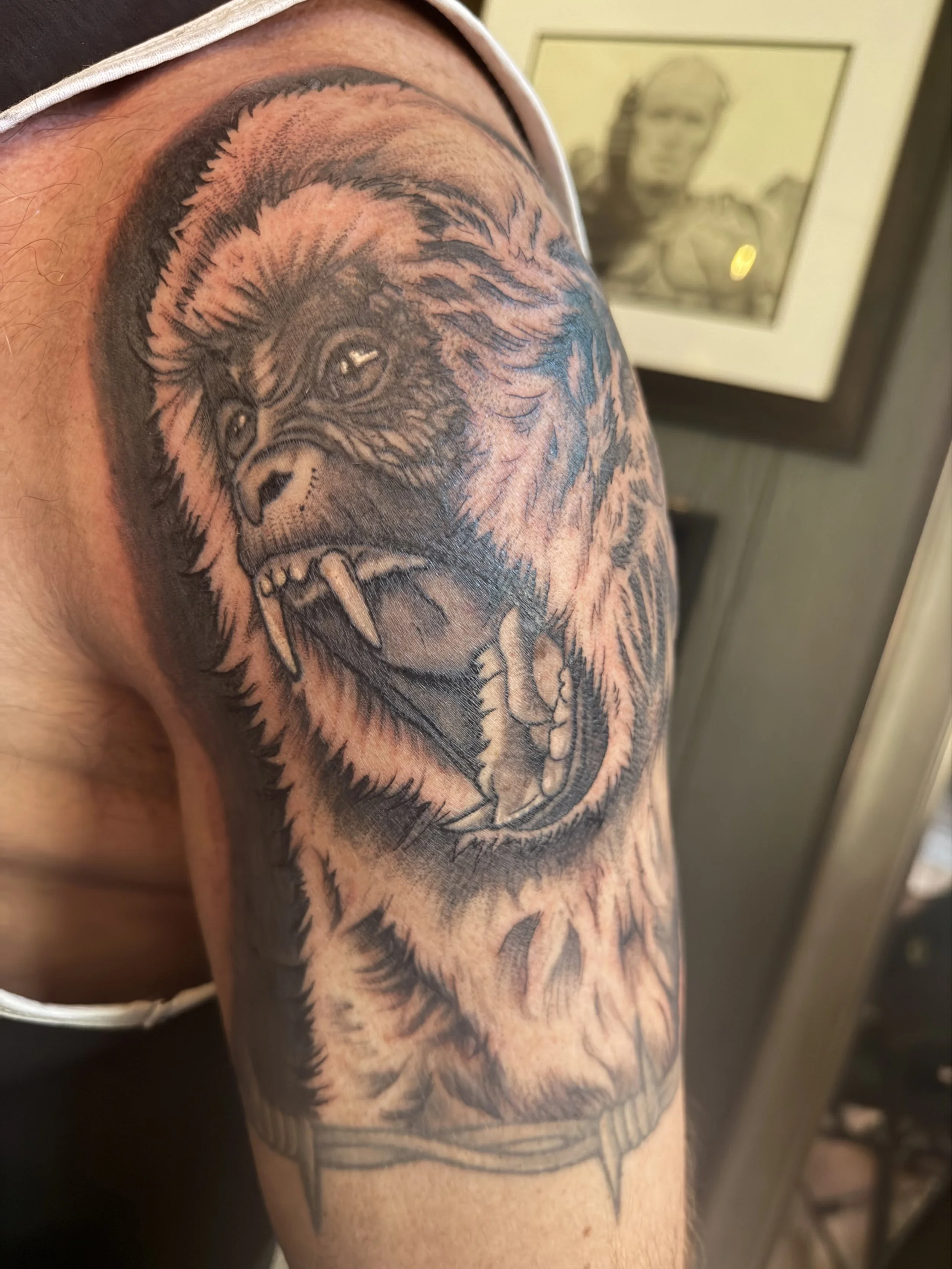 A close-up of a person's upper arm with a detailed black and gray tattoo of a roaring bear's head, showing its open mouth, sharp teeth, and intense eyes.
