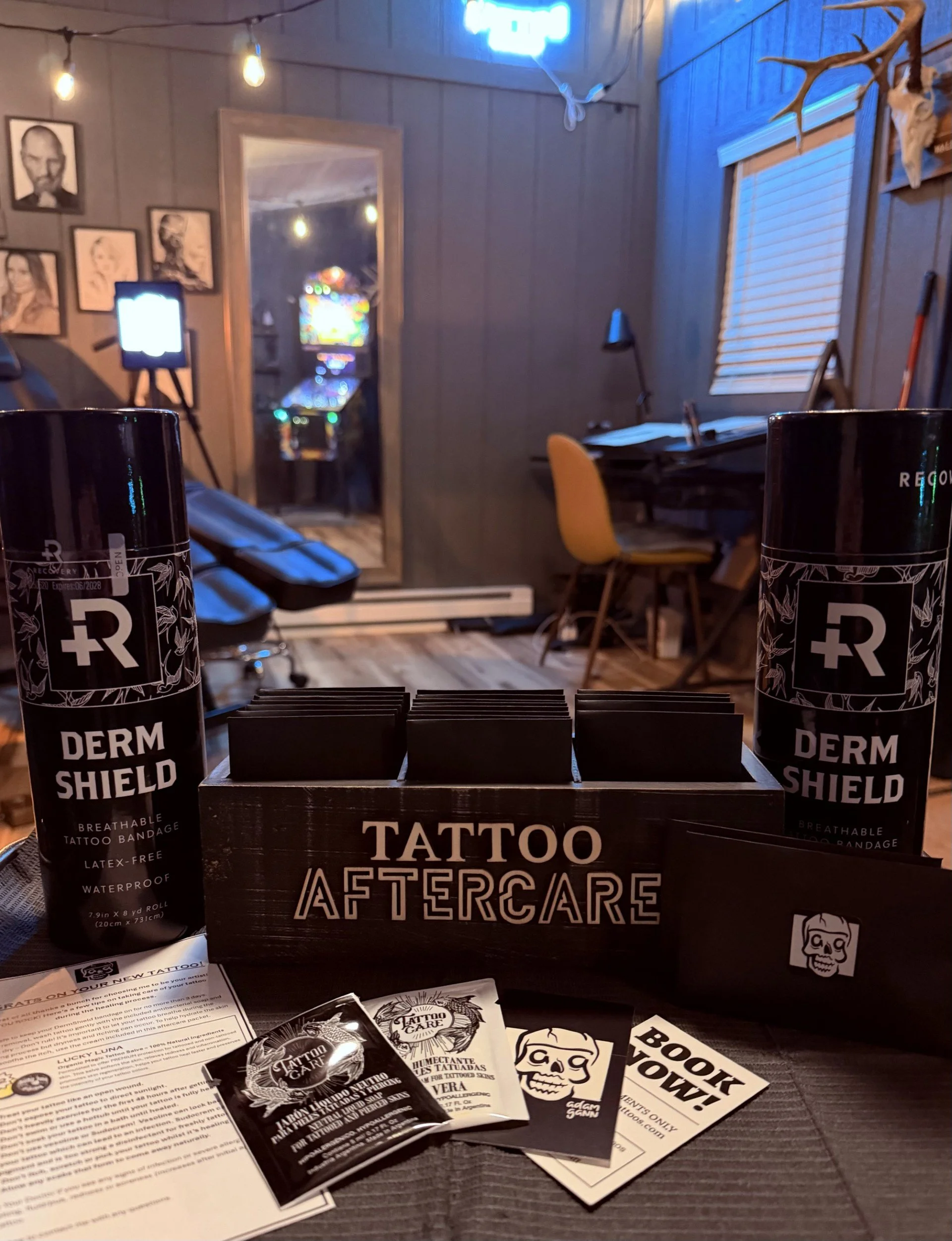 A tattoo aftercare display includes a black box labeled 'Tattoo Aftercare,' two cans labeled 'Derm Shield' spray, pamphlets and stickers with tattoo-themed artwork, and some informational papers on a dark wooden surface. The background shows a room with framed art portraits, string lights, a mirror, a window with blinds, and a desk with a yellow chair.