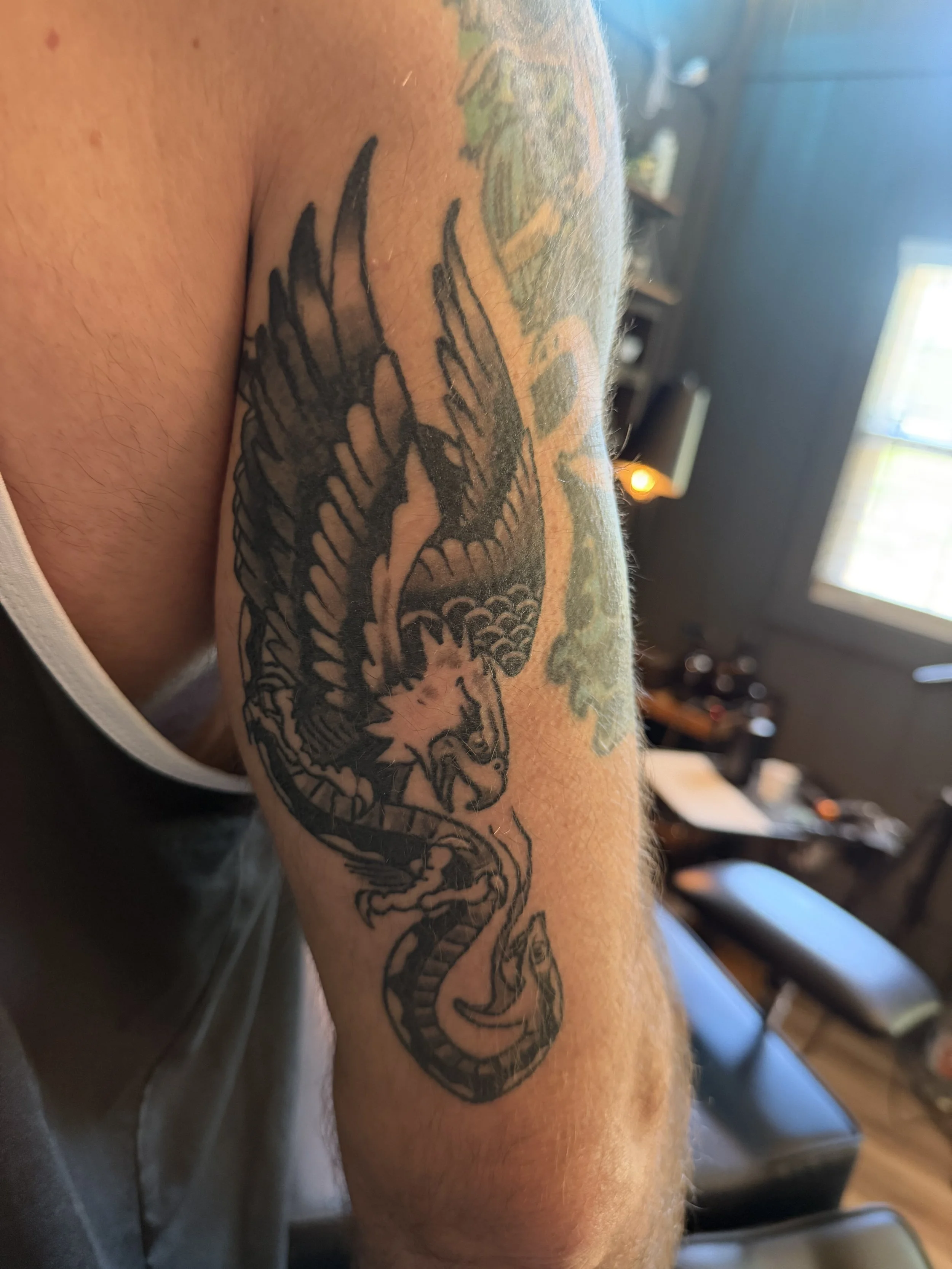 A tattoo of a dragon on a person's upper arm, with a background of a room with a window and furniture.