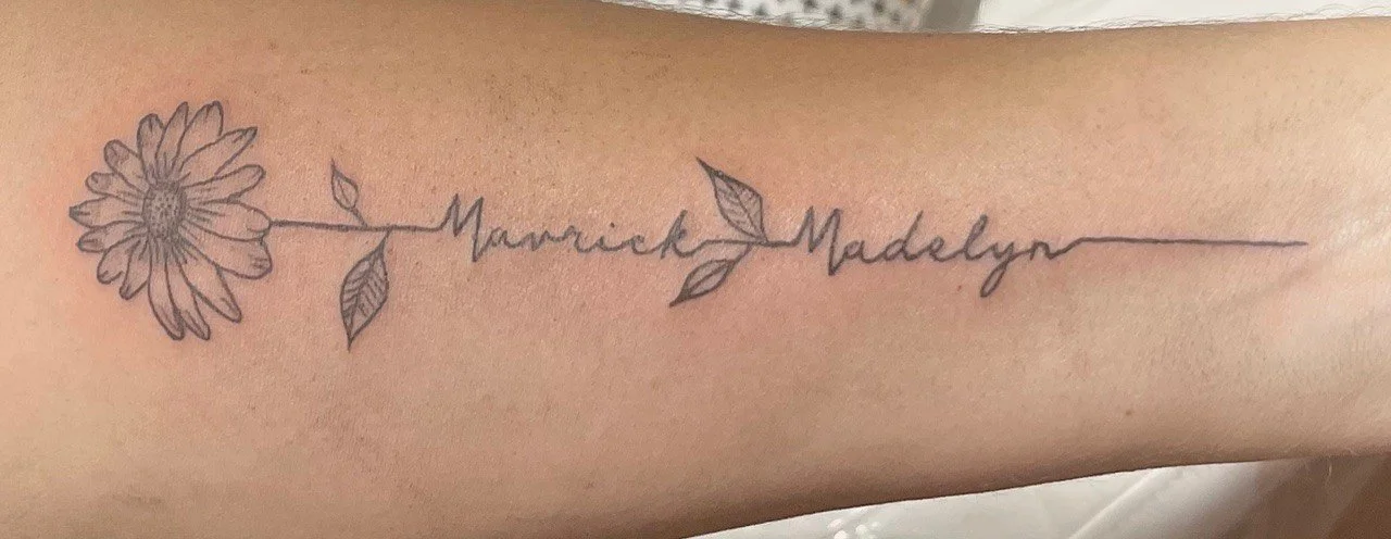 Tattoo of a daisy flower connected to cursive text that reads 'Maverick Madelyn' on a person's arm.