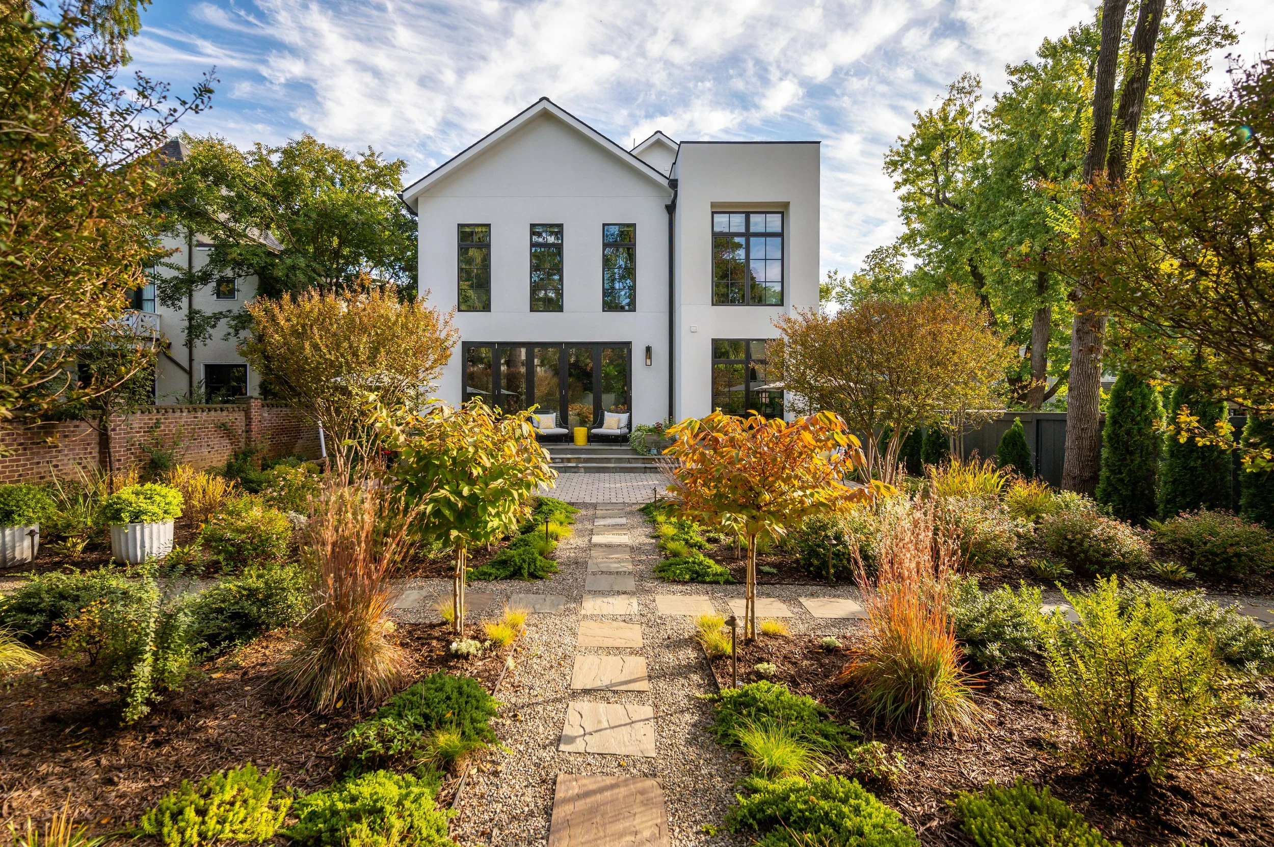 Modern white house with large windows, front porch with seating, surrounded by a landscaped garden with trees and plants, under a partly cloudy sky.