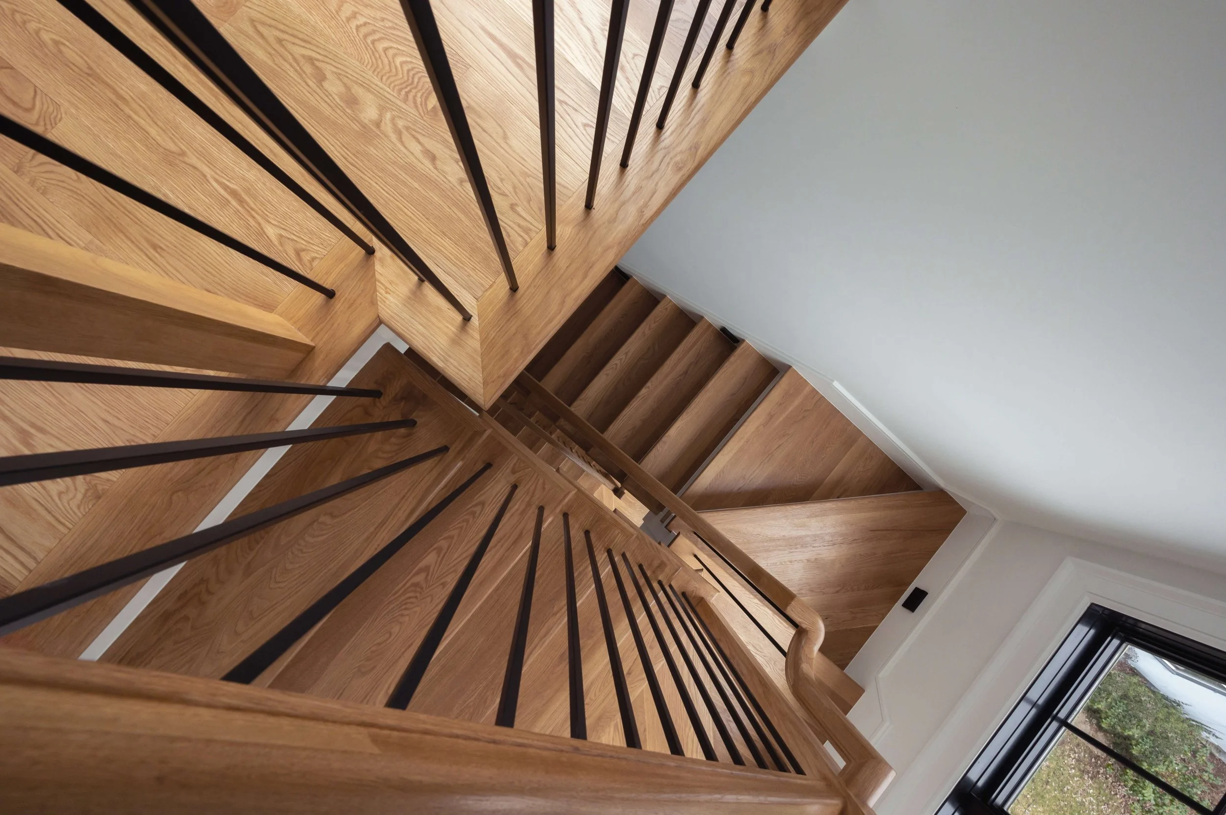 View looking down a wooden staircase with black metal balusters and a wooden handrail, next to a white wall and a window showing outdoor greenery.