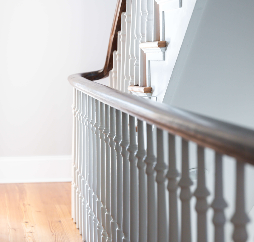 Inside view of a staircase with a wooden handrail and white spindles, leading to the upper floor of a house. The wall and floor are light-colored.