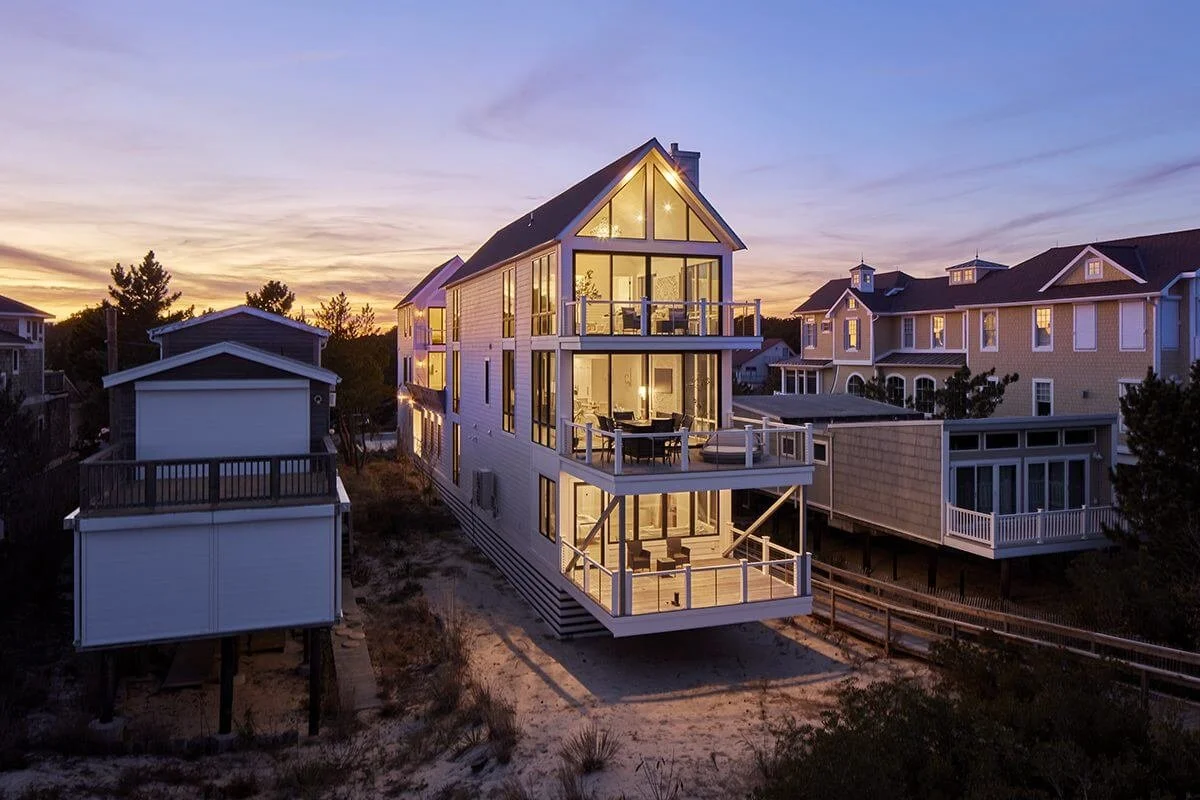 A modern multi-story house with large windows and balconies, lit from within, situated on a sandy lot during sunset with neighboring houses nearby.