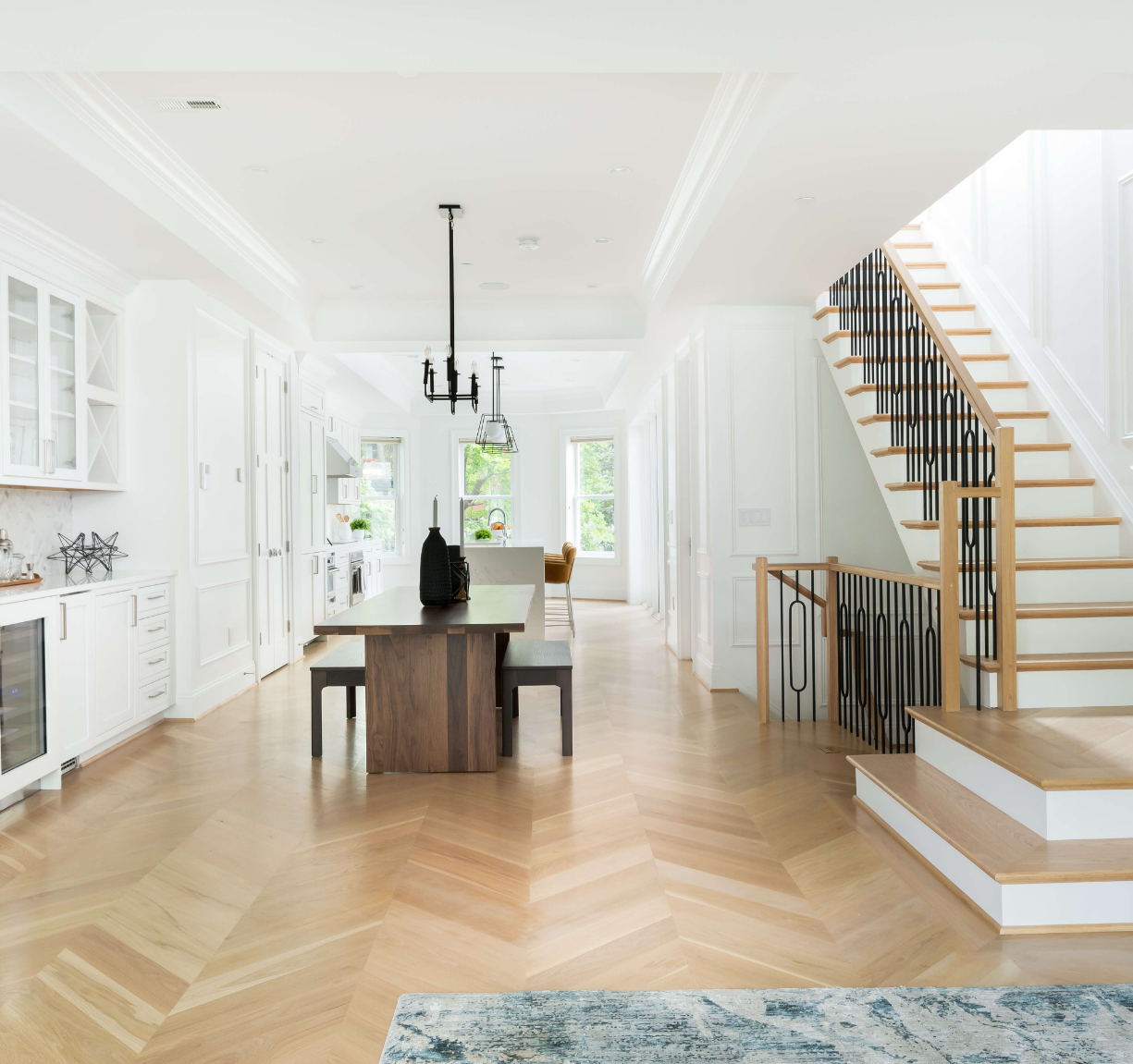 Bright open-concept kitchen and living area with light wood flooring, white walls, a staircase with black railings, a kitchen island, and large windows with greenery outside.