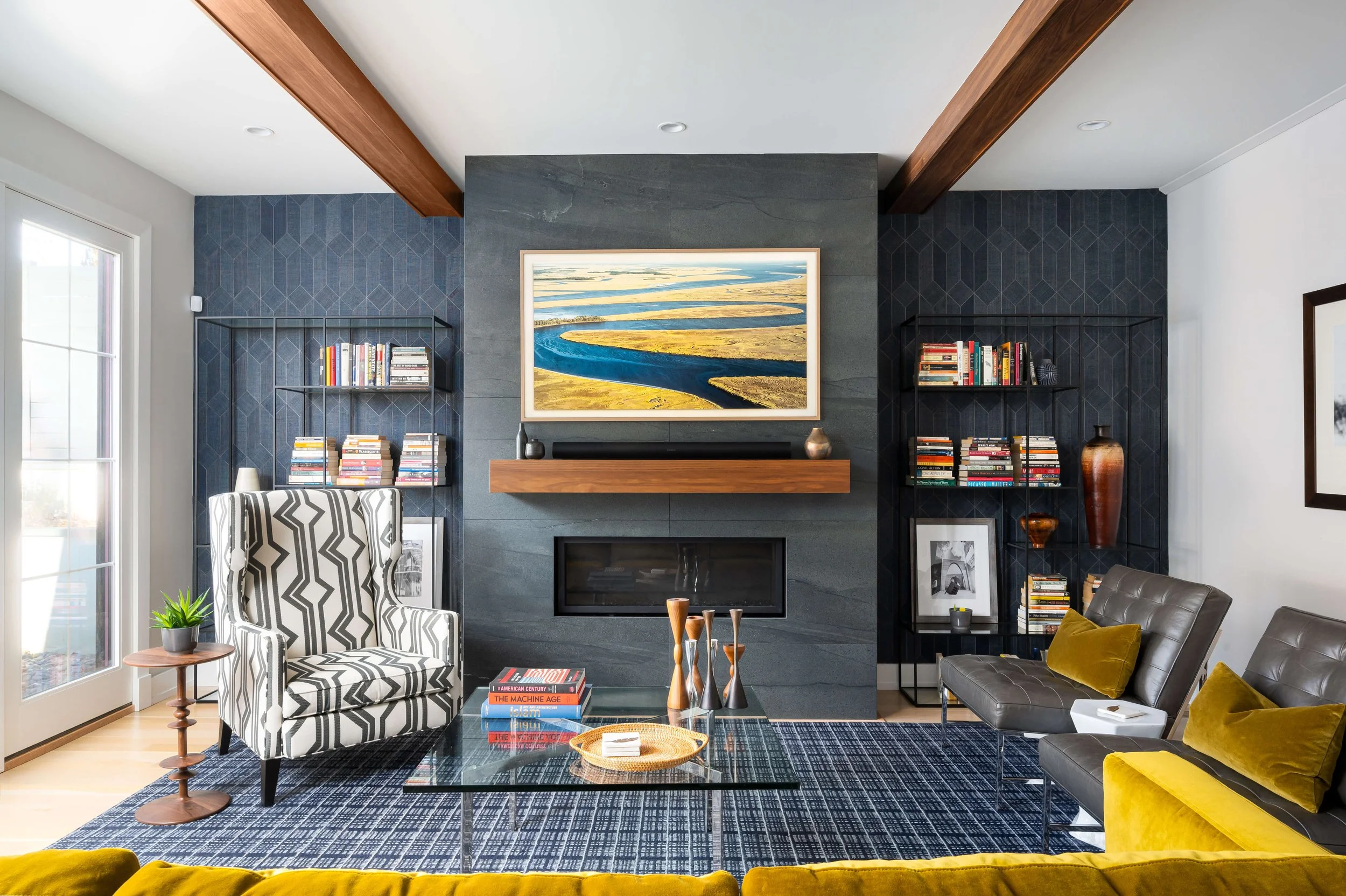 Living room with a fireplace, a large colorful landscape painting above it, black built-in bookshelves with books and decor, a patterned armchair, a glass coffee table with books and decorative items, yellow velvet pillows, a rug with a blue pattern, a side table with a plant, and gray chairs in a modern style.