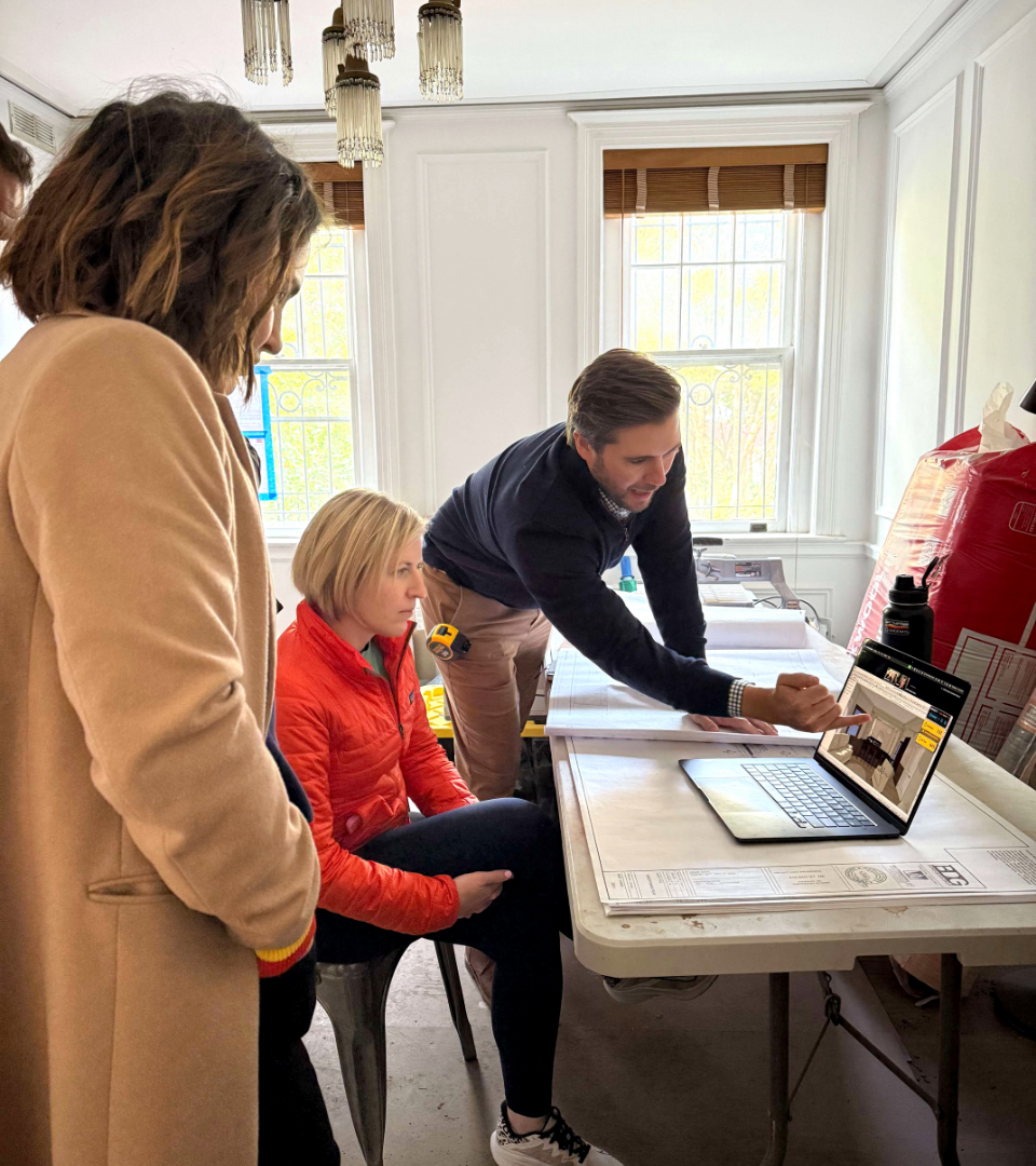 Three people in a room looking at a laptop on a table, with two women standing and a woman sitting.