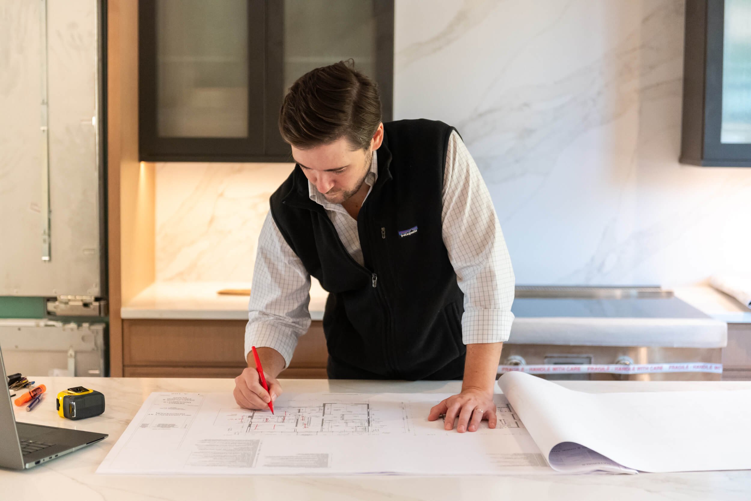 Man reviewing architectural plans on a kitchen island with laptop and tools nearby.