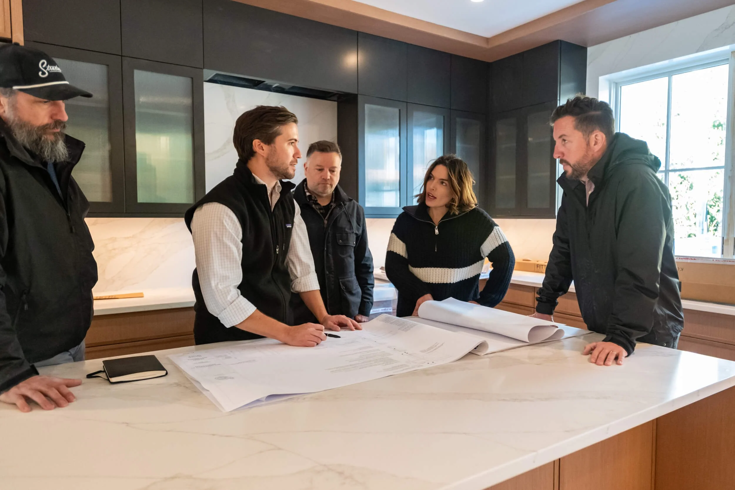 Five people are gathered around a kitchen island with blueprints, having a discussion.
